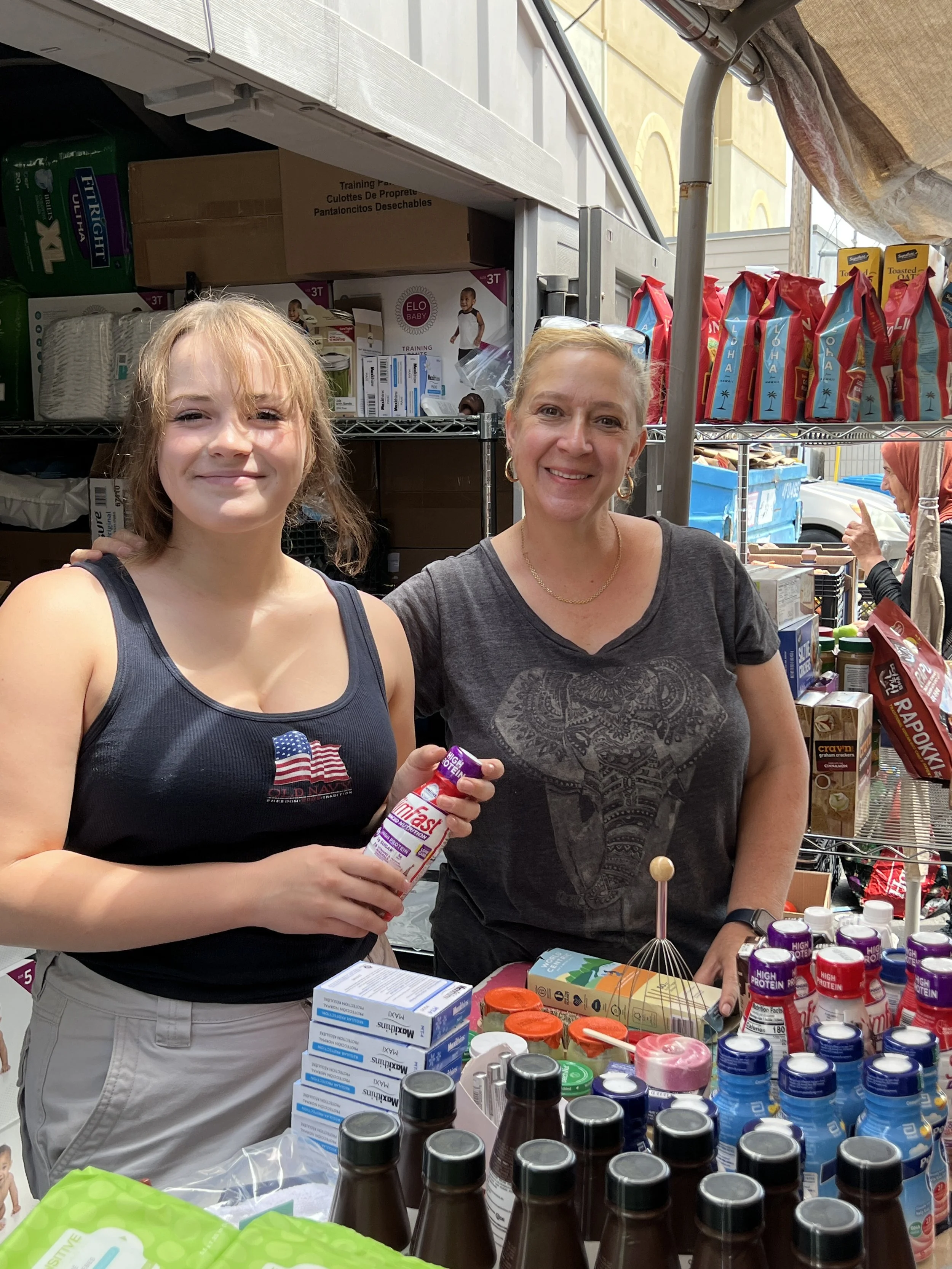 Two women smiling inside a store, surrounded by shelves with various health and food products, some of which include bottles of supplements or vitamins, boxes, and packaged snacks.