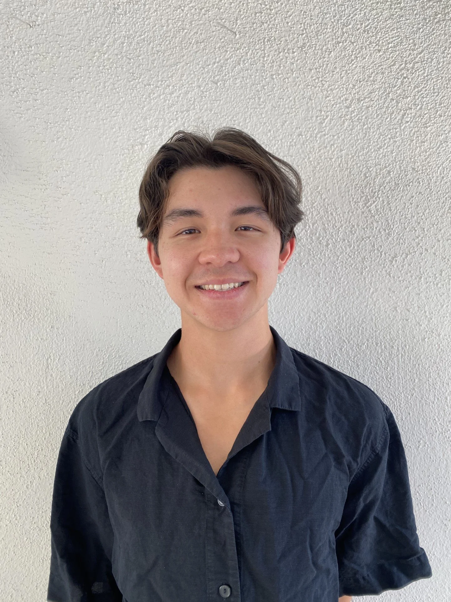 A young man smiling in front of a plain off-white wall, wearing a black button-up shirt with the top button undone.