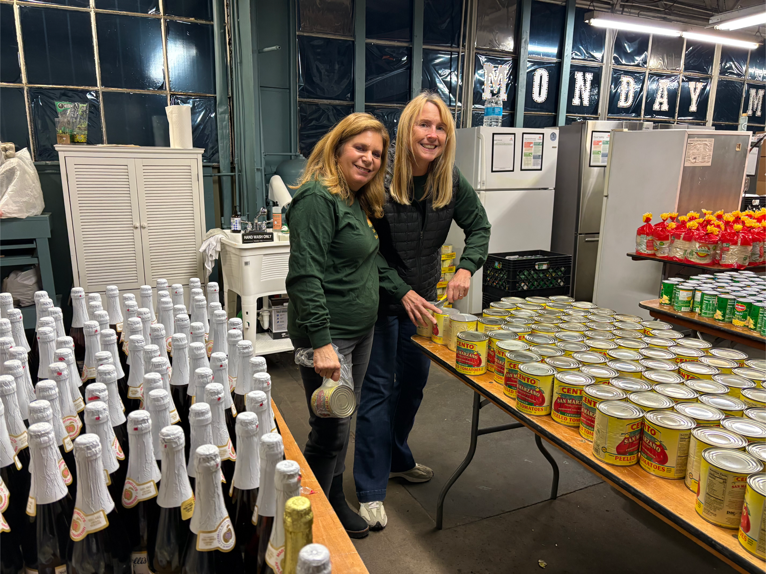 Two women standing behind tables filled with canned vegetables, bottled beverages, and bottled sparkling drinks in a warehouse-like setting with decorations that spell out 'Monday' in the background.
