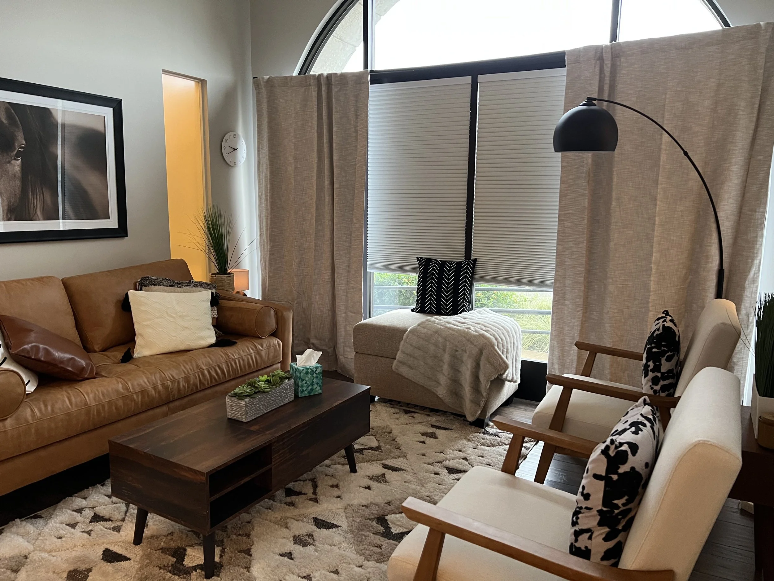 Living room with tan leather sofa, beige armchair with black and white patterned pillows, window with beige curtains, wall art, indoor plants, coffee table, and area rug.