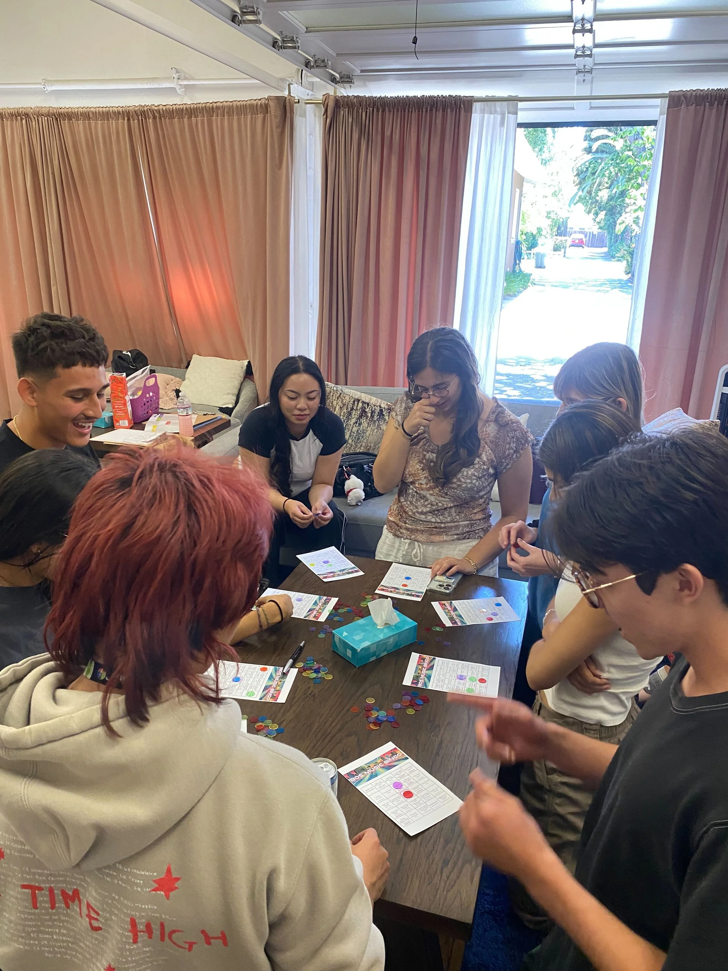 Group of people playing a board game around a table in a living room with pink curtains, a couch, and an open door showing a sunny outdoor scene.