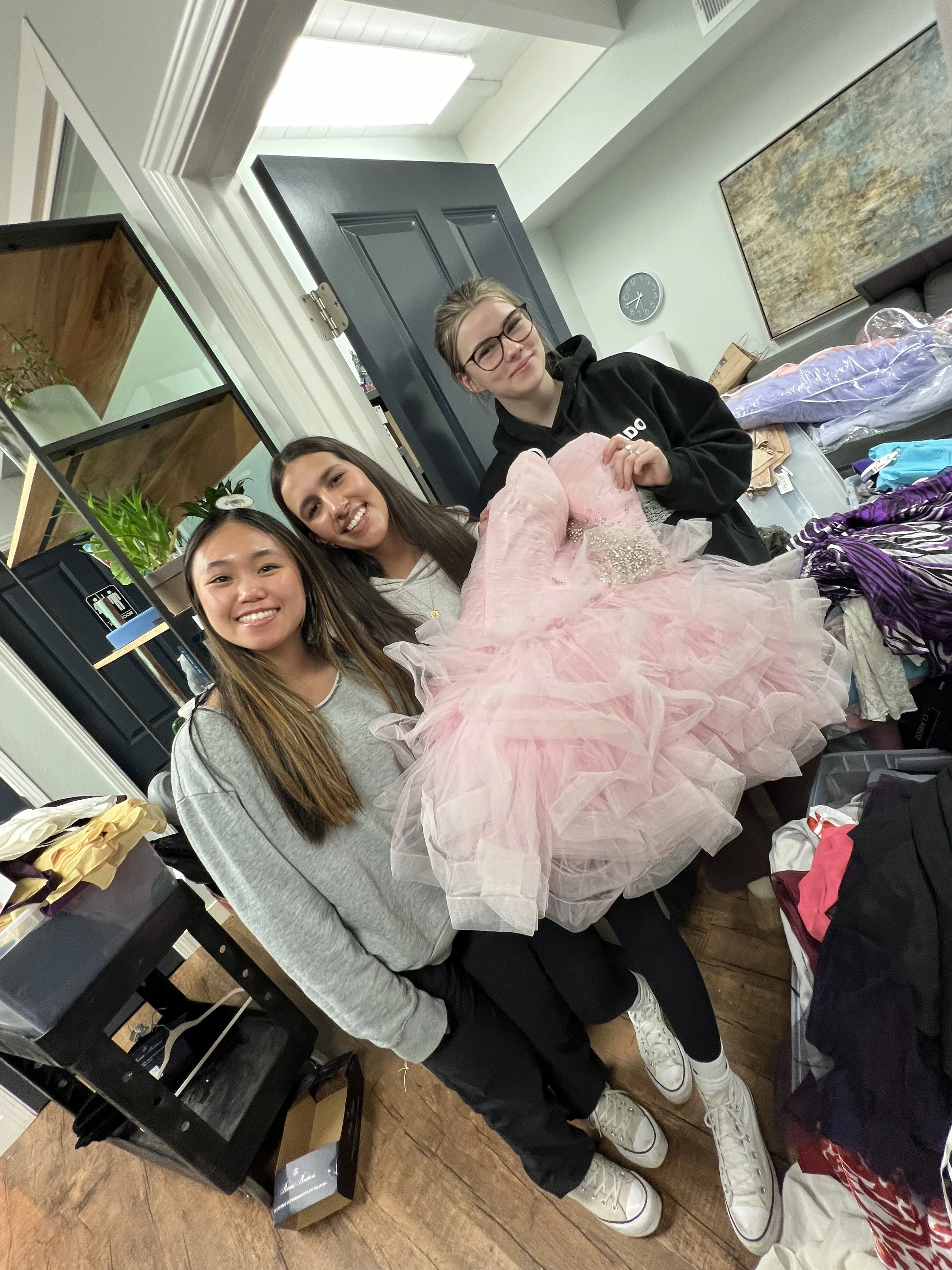 Three young women in casual clothing are sitting at a table with a pink, ruffled dress in front of them, in a room with shelves, plants, and a clock on the wall.
