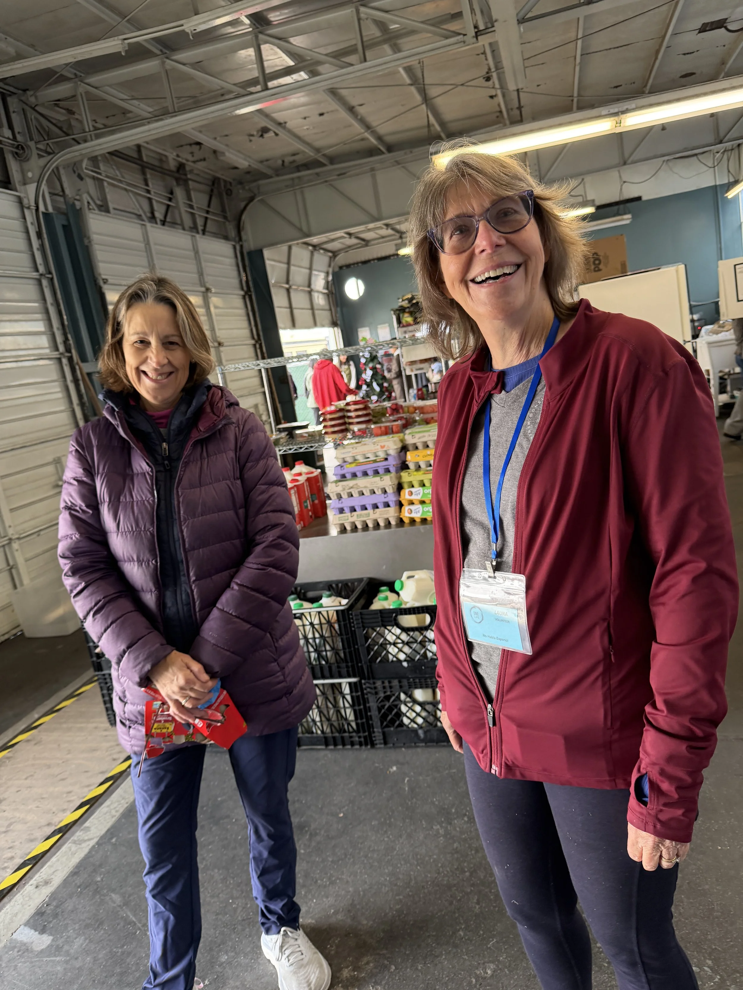Two women standing inside a warehouse with tables of food items behind them, smiling at the camera.
