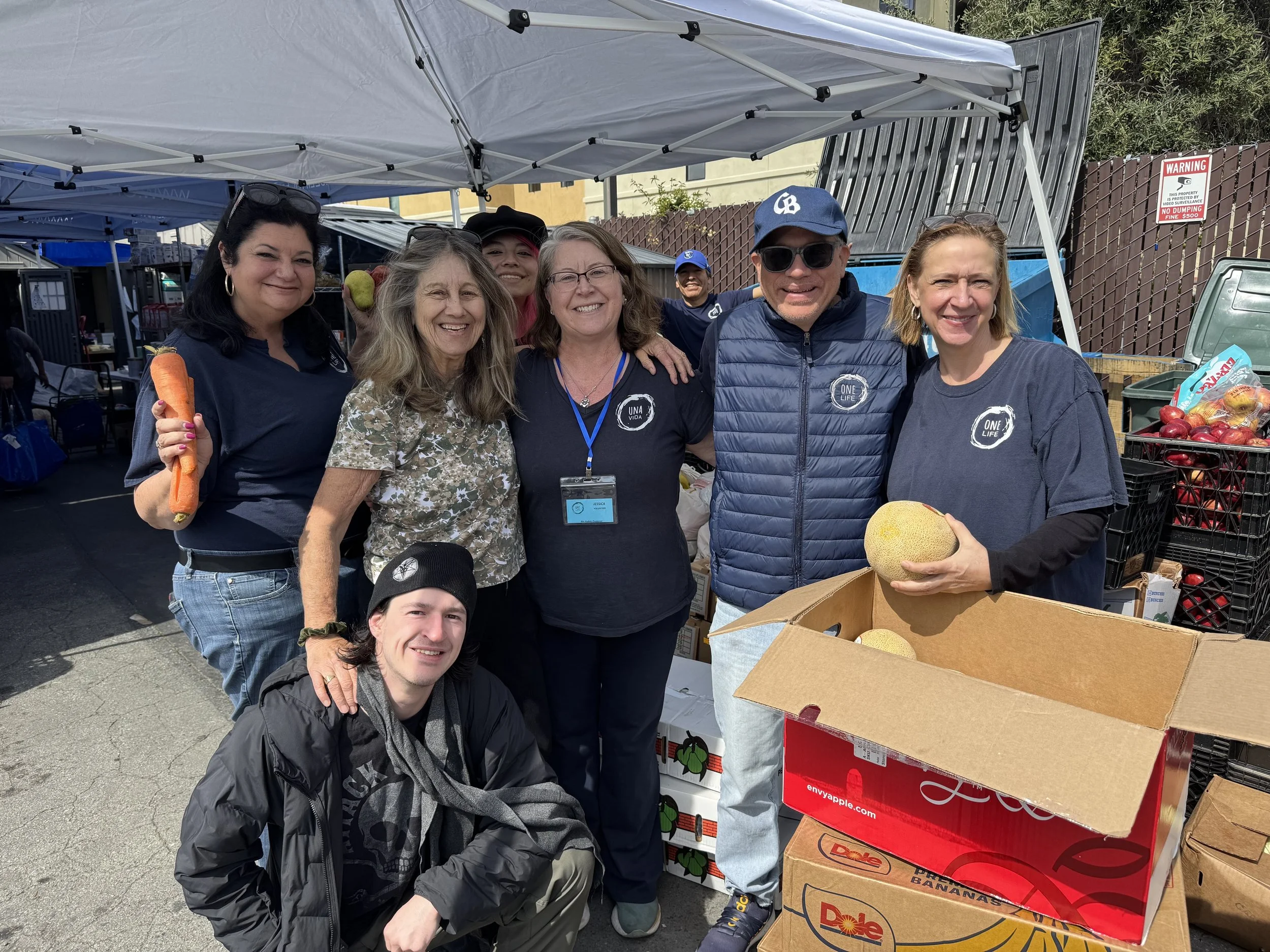 A group of seven people smiling and standing together at a farmers market stall, holding fruits and vegetables, with boxes of produce including bananas and melons in front of them.