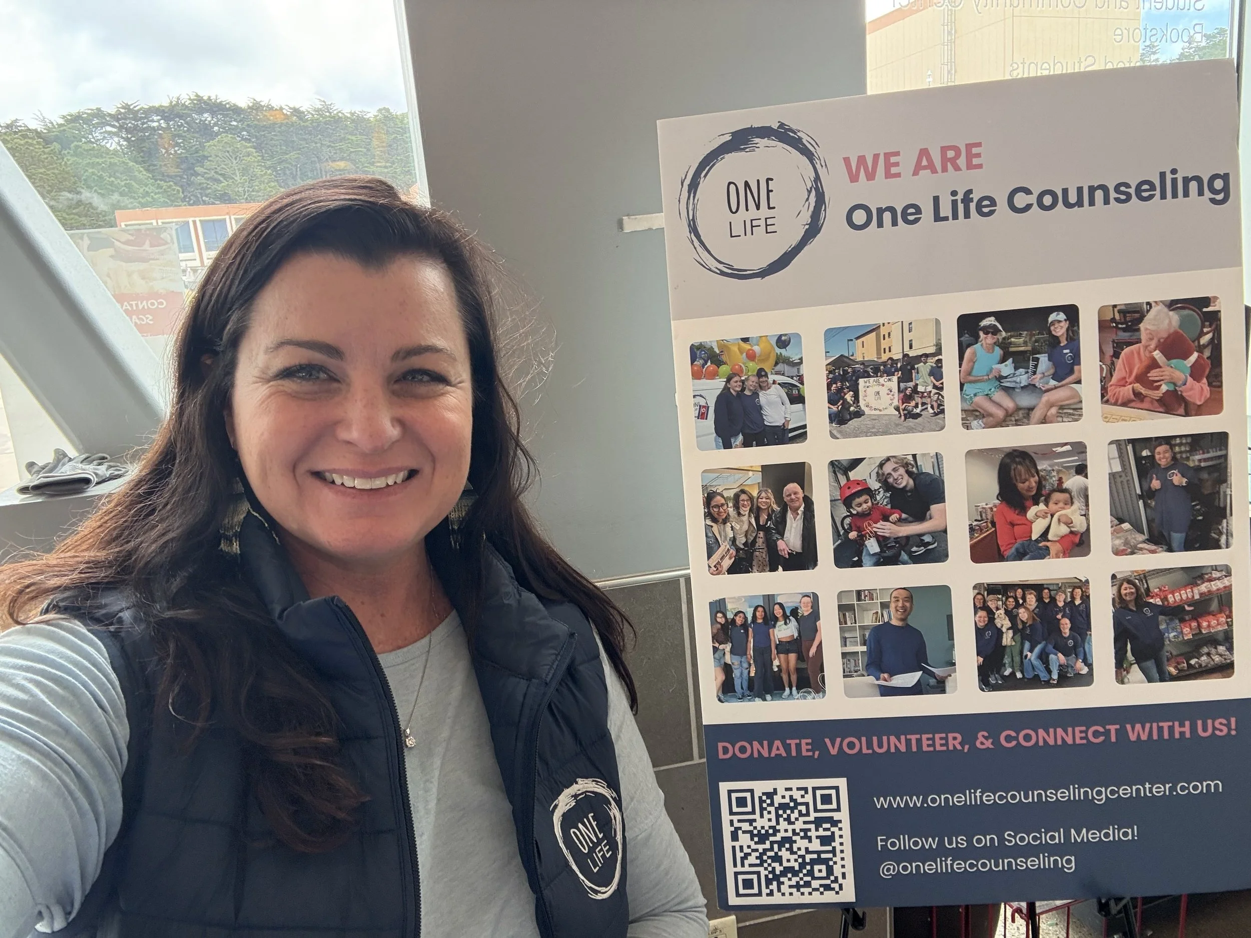 Woman smiling at the camera next to an informational display board promoting One Life Counseling with photos of community events and activities, social media links, and a QR code.