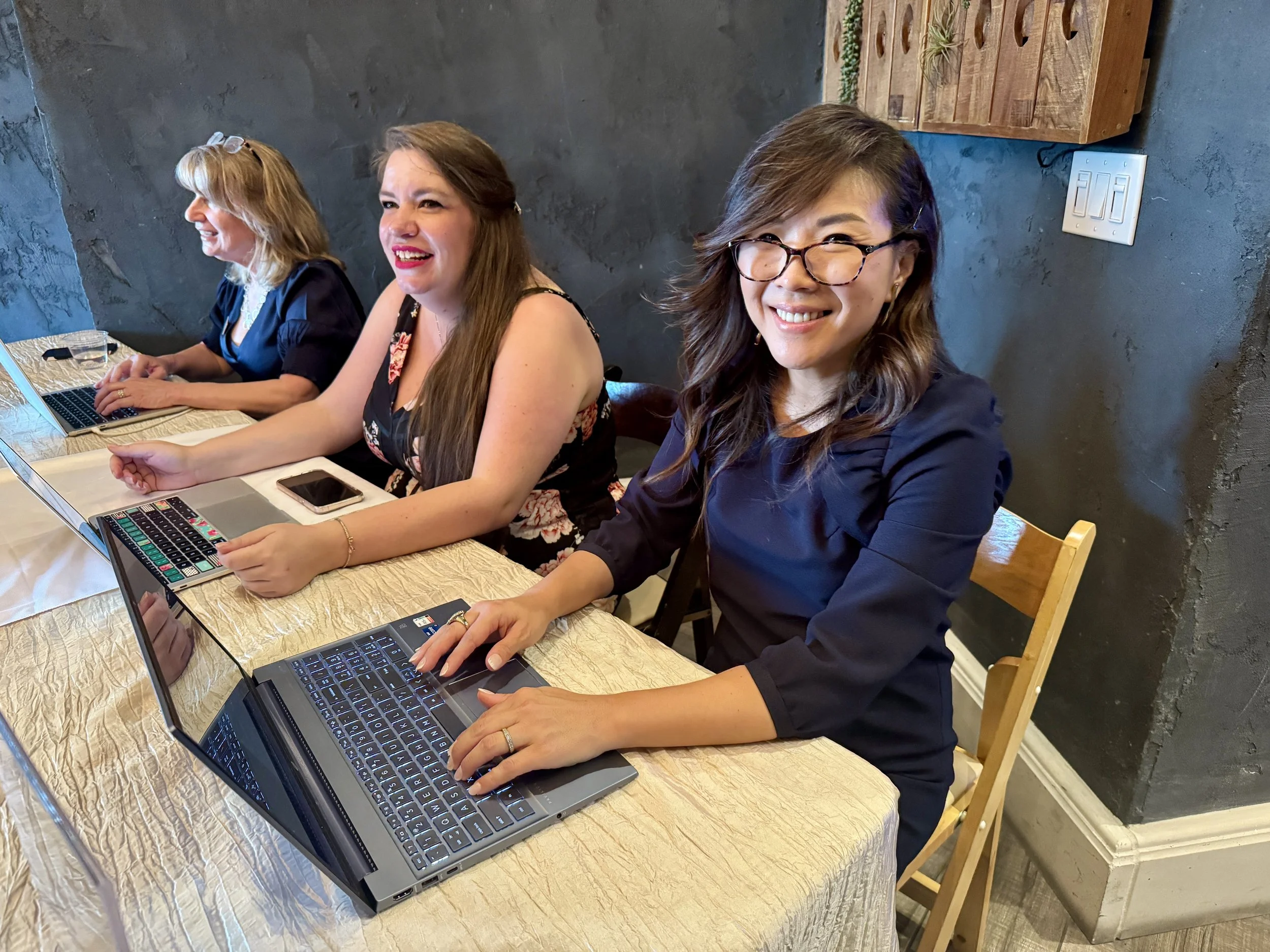 Three women sitting at a table with laptops, smiling, in a room with dark textured walls.
