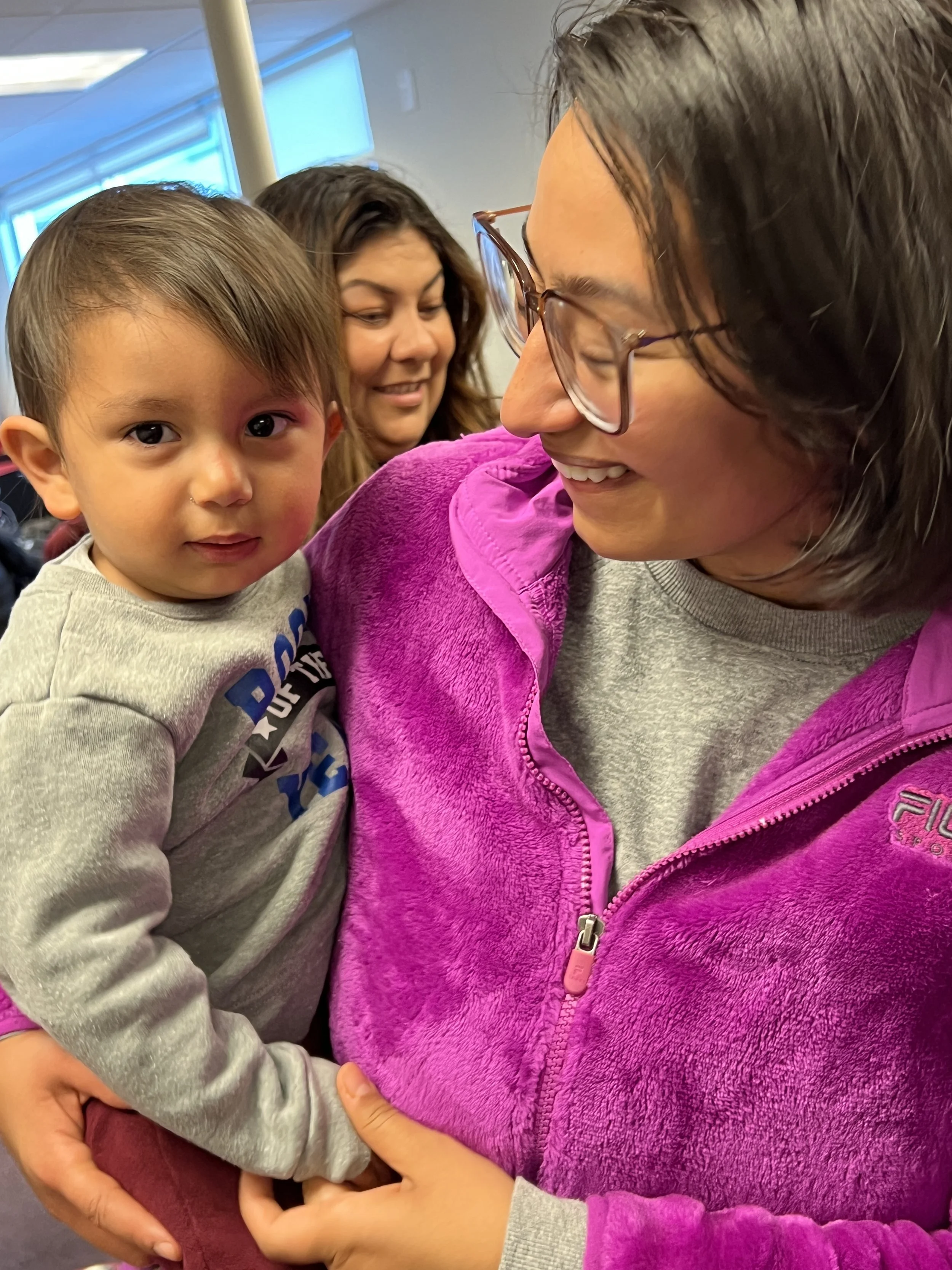A woman smiling and holding a young boy close while another woman smiles behind them in the background.