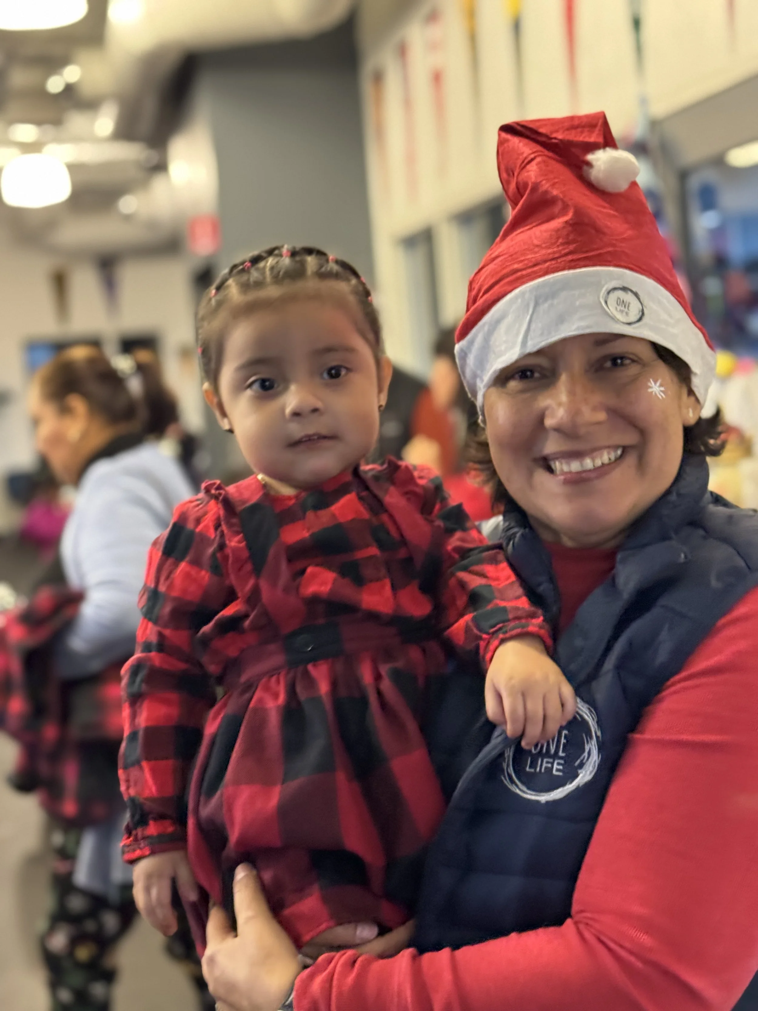 A woman holding a young girl at a festive event, both dressed in Christmas-themed attire. The woman is wearing a Santa hat and a vest with a logo, smiling at the camera, while the girl wears a red and black checkered dress and has braided hair.