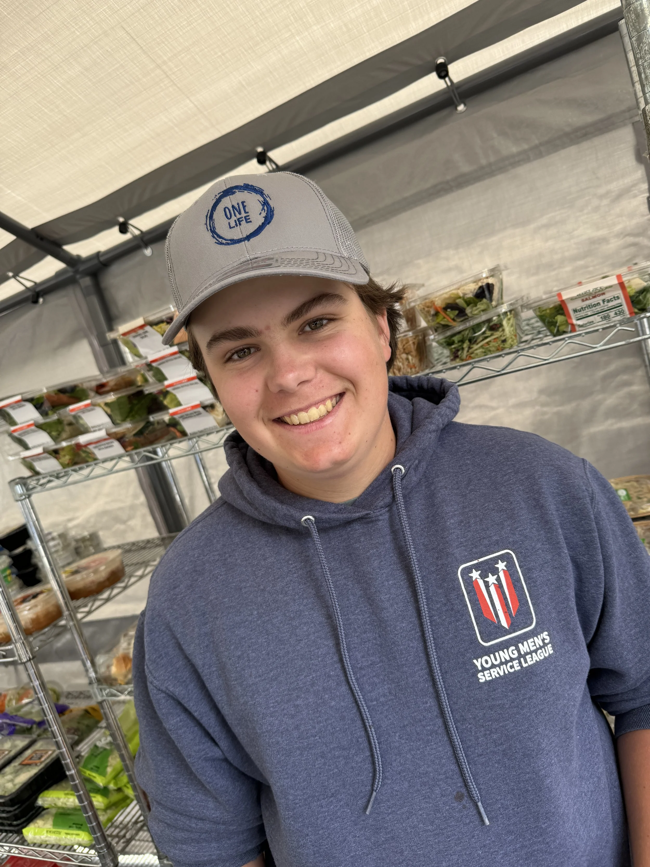 Smiling young man wearing a gray hoodie with the Young Men's Service League logo and a gray cap that says One Life, standing in front of metal shelves filled with pre-packaged salads and food items.