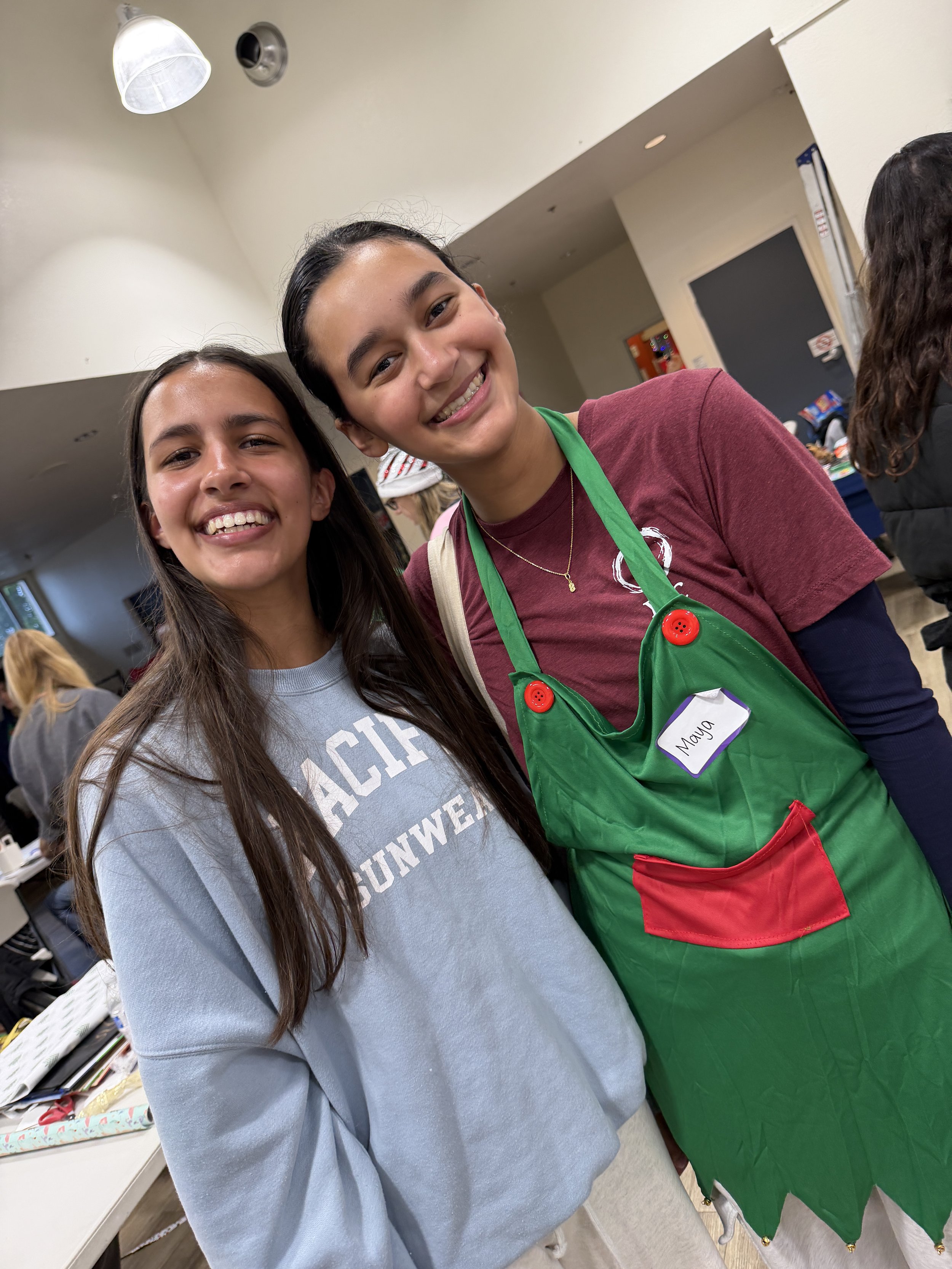 Two young women smiling at a camera, one wearing a gray sweatshirt and the other in a maroon shirt with a green apron and a name tag that says 'Mayra'.