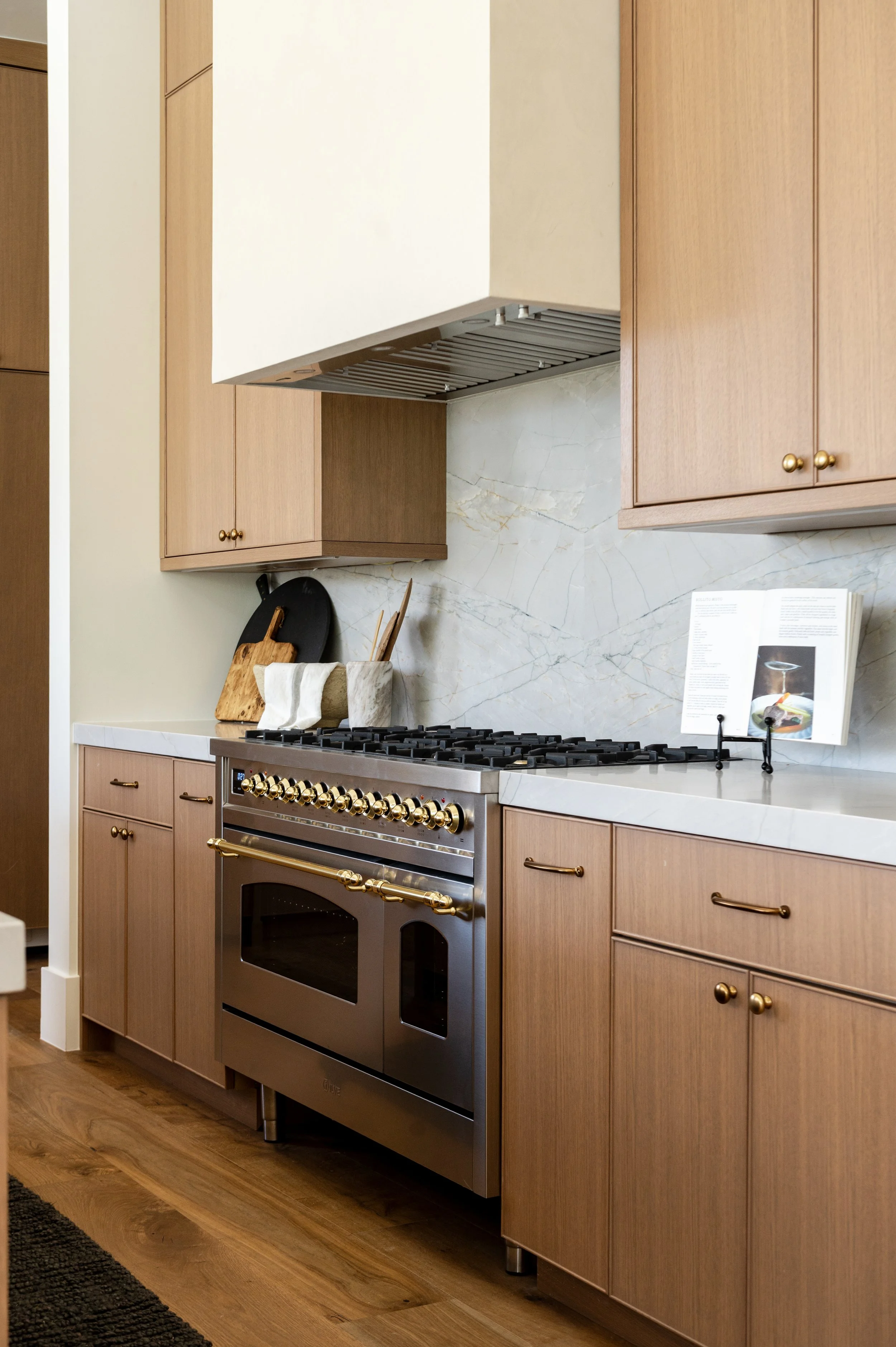 A kitchen with wooden cabinets and a stainless steel oven stove, white marble countertop, and a marble backsplash. There are cutting boards and an open cookbook on a stand.