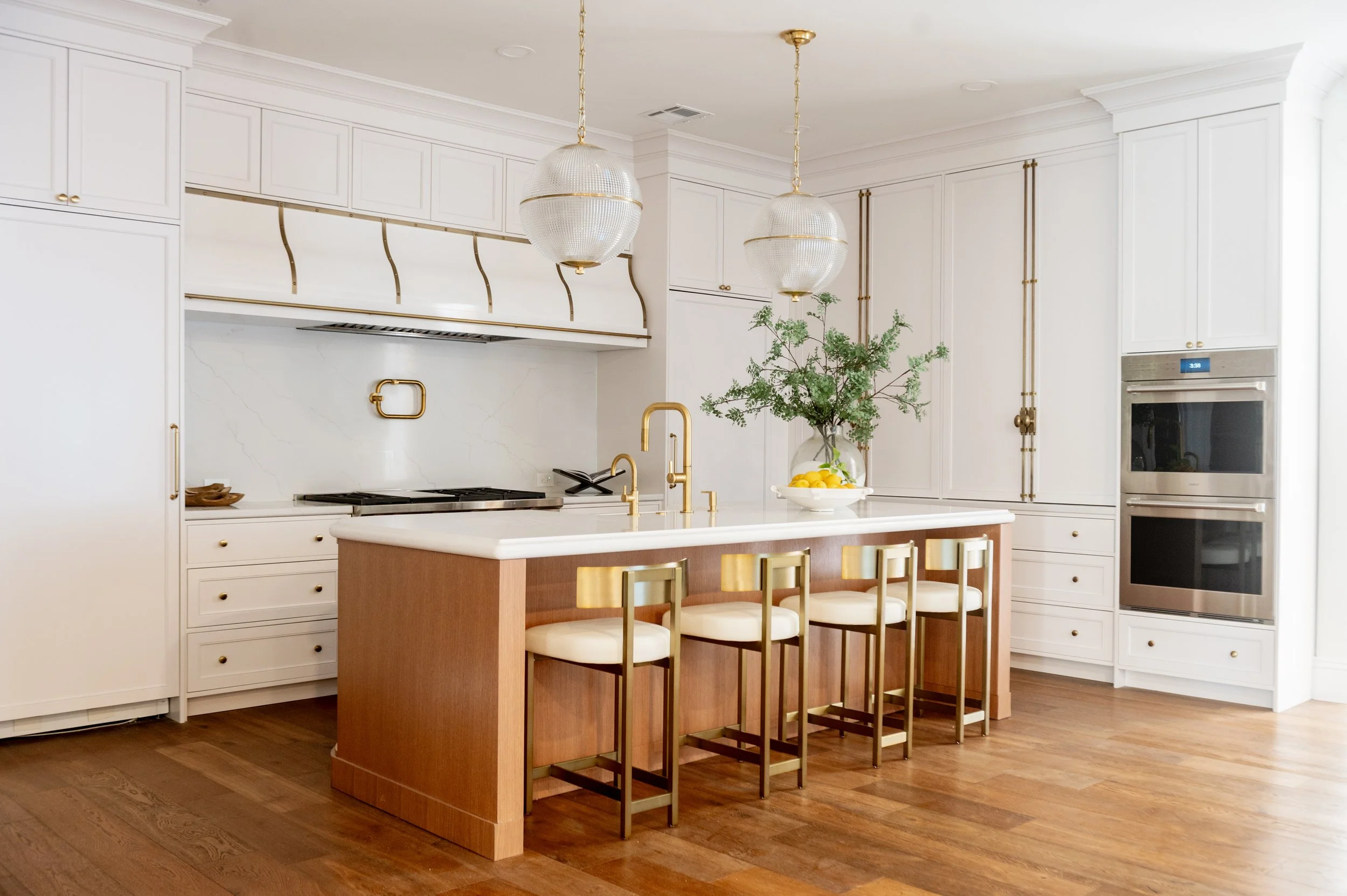 A modern kitchen with white cabinets, a central island with a wood base and white countertop, and four matching bar stools with gold frames and white cushions. Two large pendant lights hang above the island, and there is a vase with green foliage and lemons on the countertop. The kitchen features a double oven, a cooktop, and a gold faucet, with wooden flooring throughout.