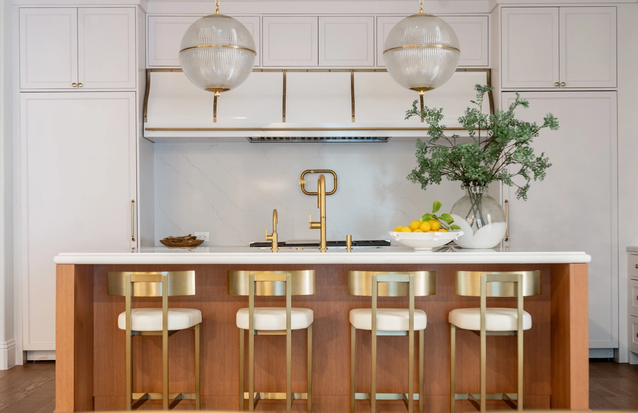 Modern kitchen with white cabinetry, a wooden island with four gold and white chairs, two large textured gold pendant lights, a marble backsplash, gold fixtures, a bowl of lemons, and a large vase with greenery.