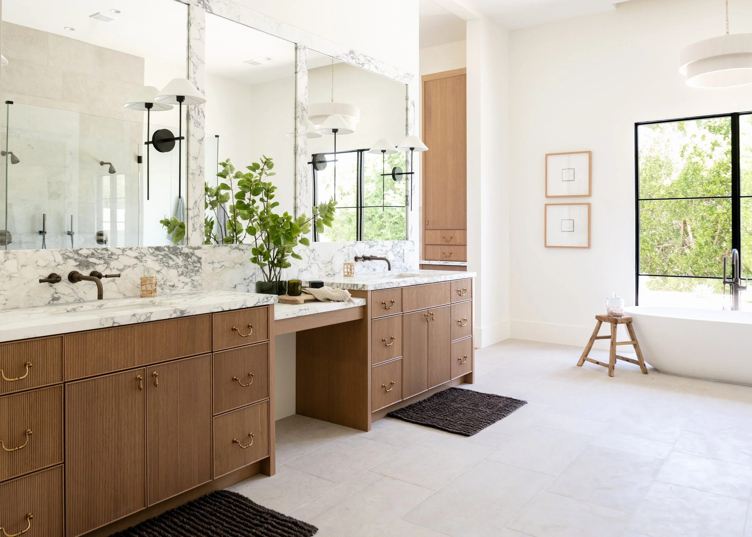 Bright bathroom with double marble countertops, wood cabinets, large mirror, sliding glass shower, bathtub near window, black fixtures, and minimal decor with green plant and framed artwork.