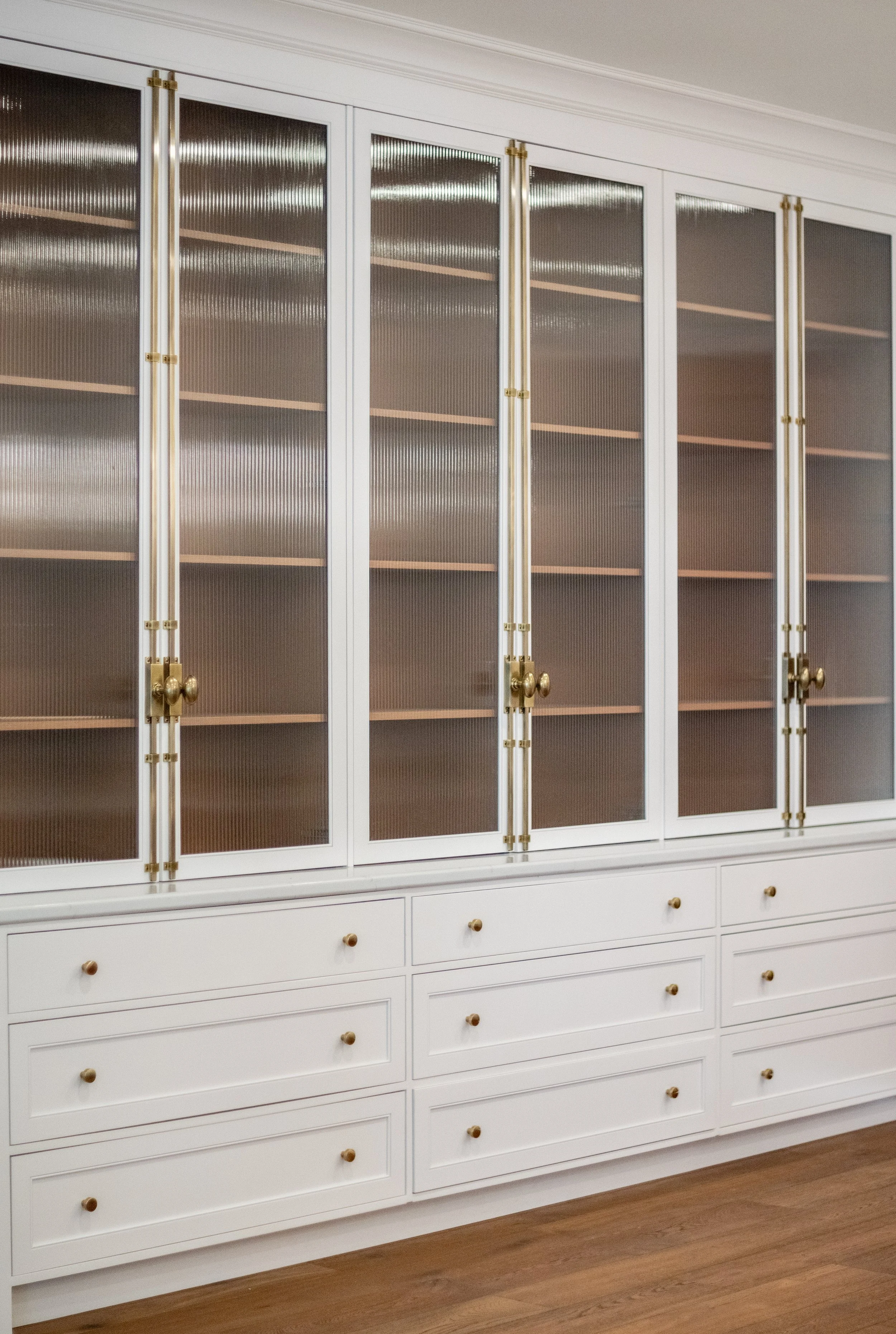 White built-in cabinet with glass doors and drawers, featuring gold hardware and handles, in a room with wooden flooring.