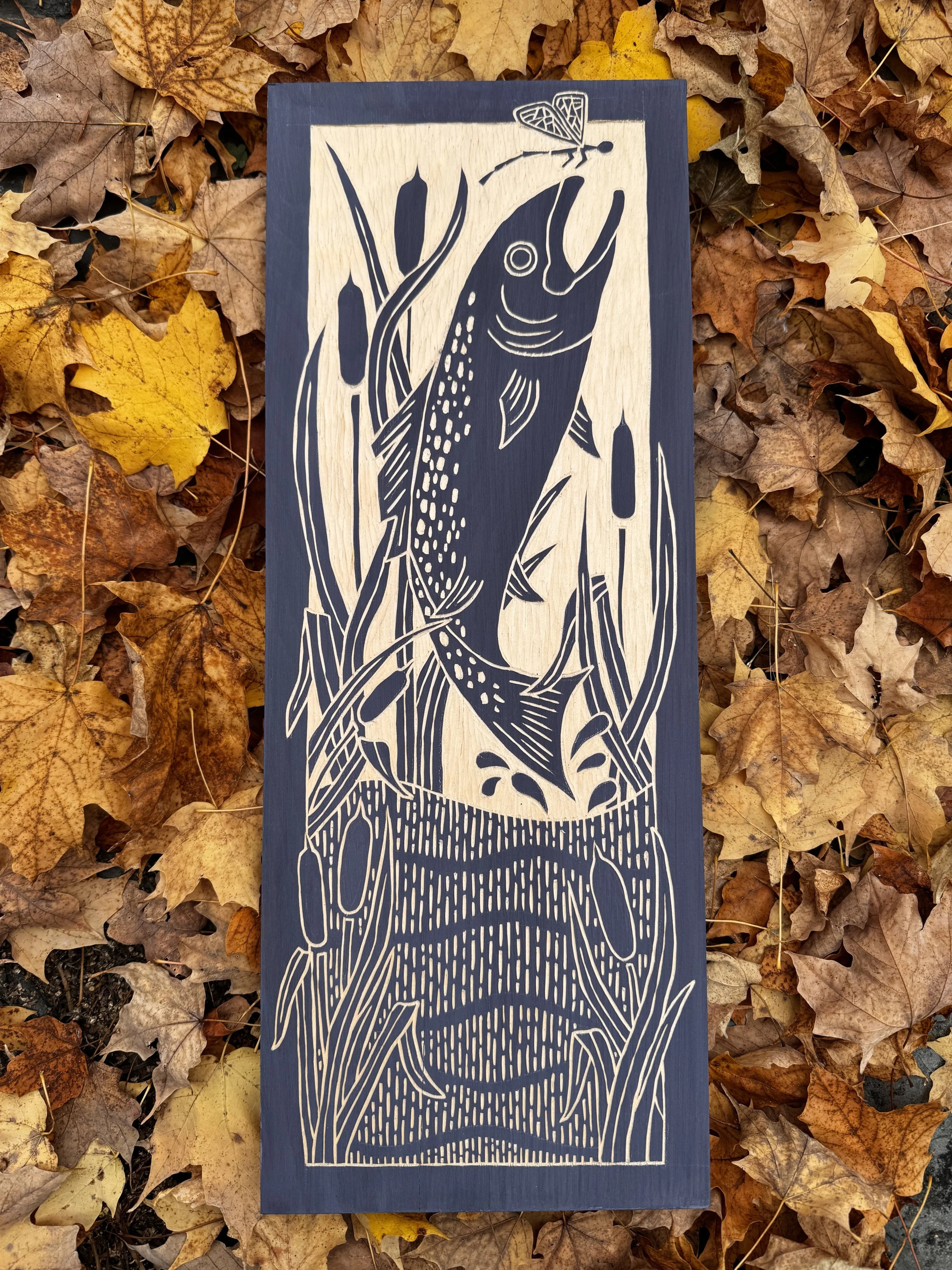 navy blue and pine hand carved design of a fish leaping out of the water with cattails and a flying insect that it appears to be trying to catch.