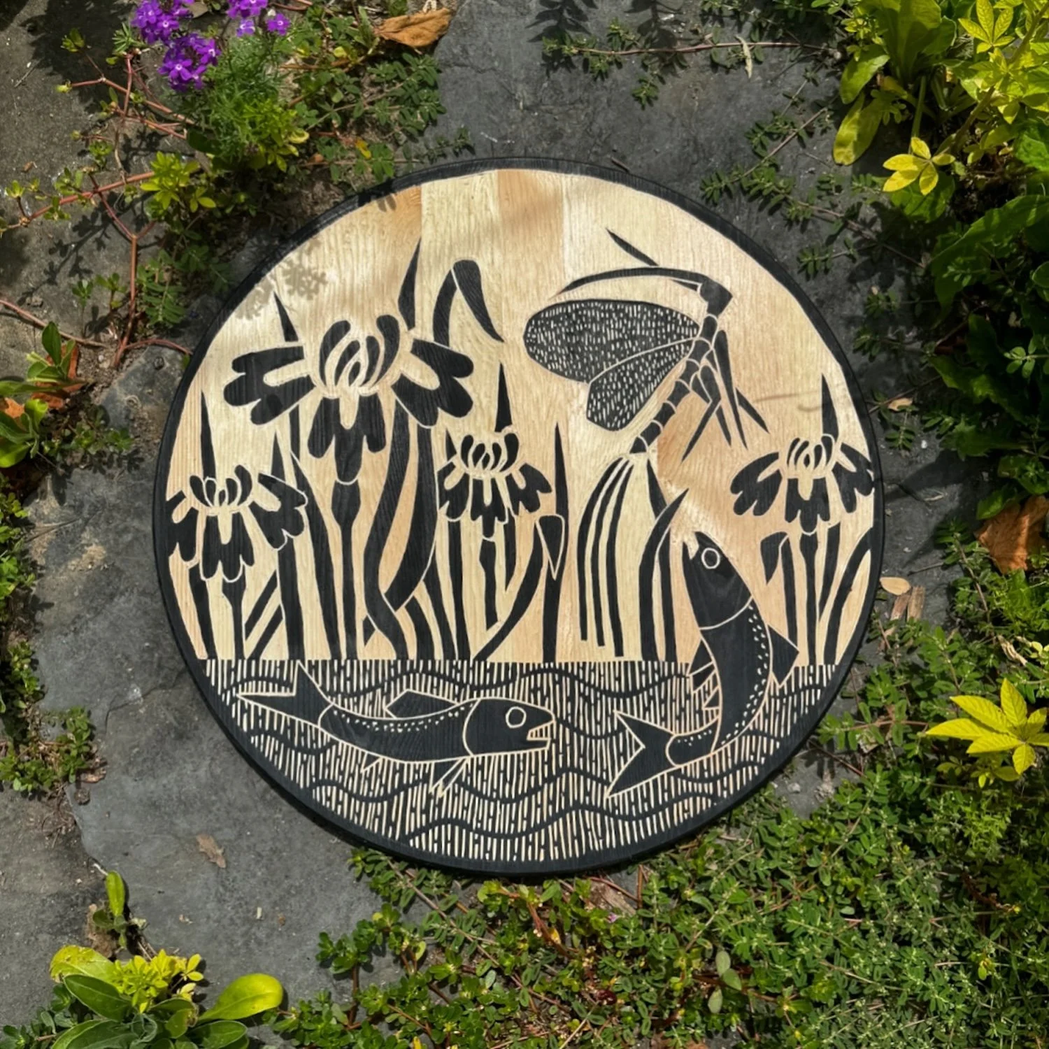 Decorative circular wooden plaque featuring black silhouette-style illustrations of flowers, a butterfly, a bee, and two fish, set on a garden stone surrounded by green plants and purple flowers.