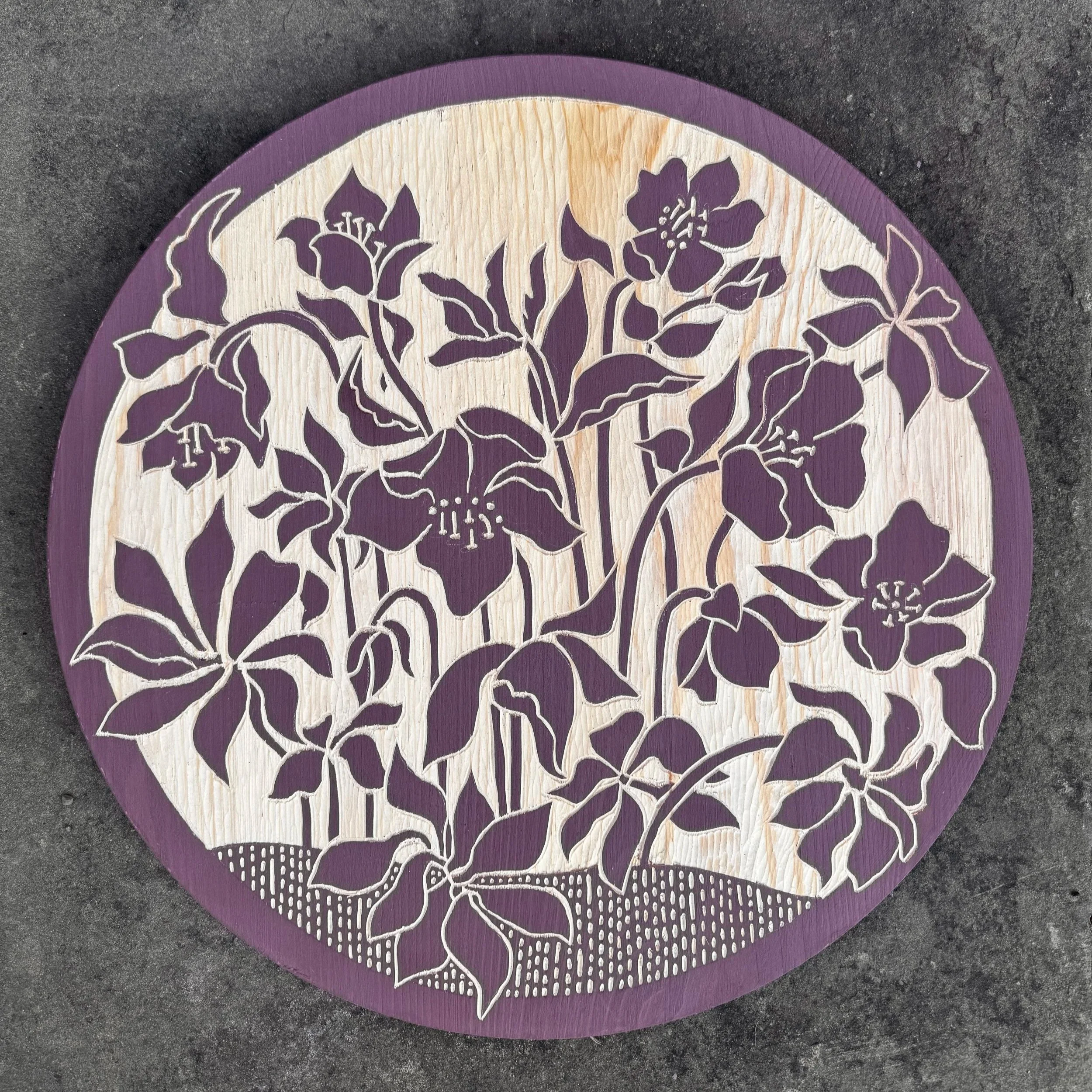 Decorative circular wood plaque with carved hellebores floral design featuring purple flowers and leaves on a light pine, with a purple border.