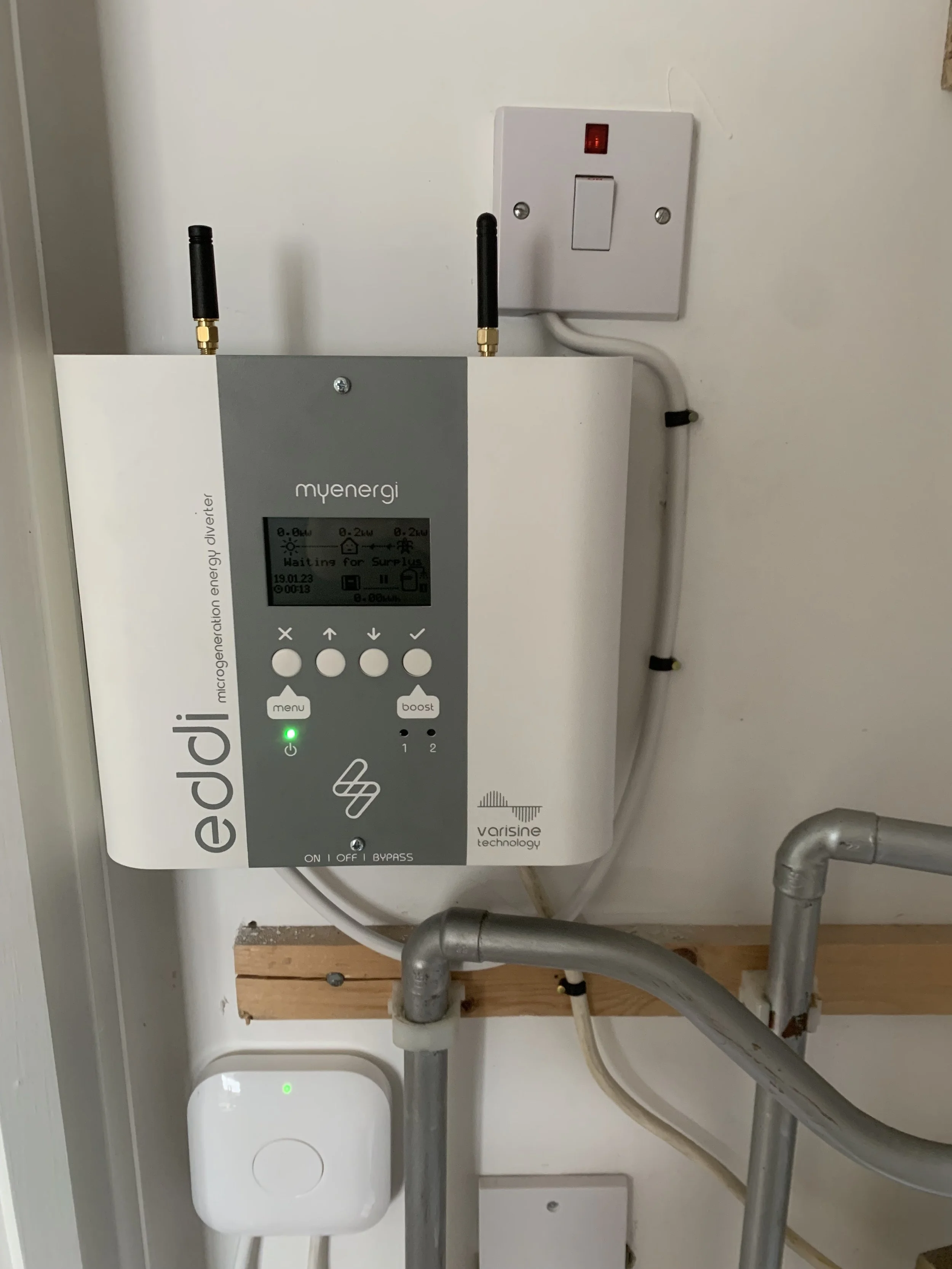 Heating controls on the wall with switch above. Silver pipes at the bottom of image above wooden panel on the wall