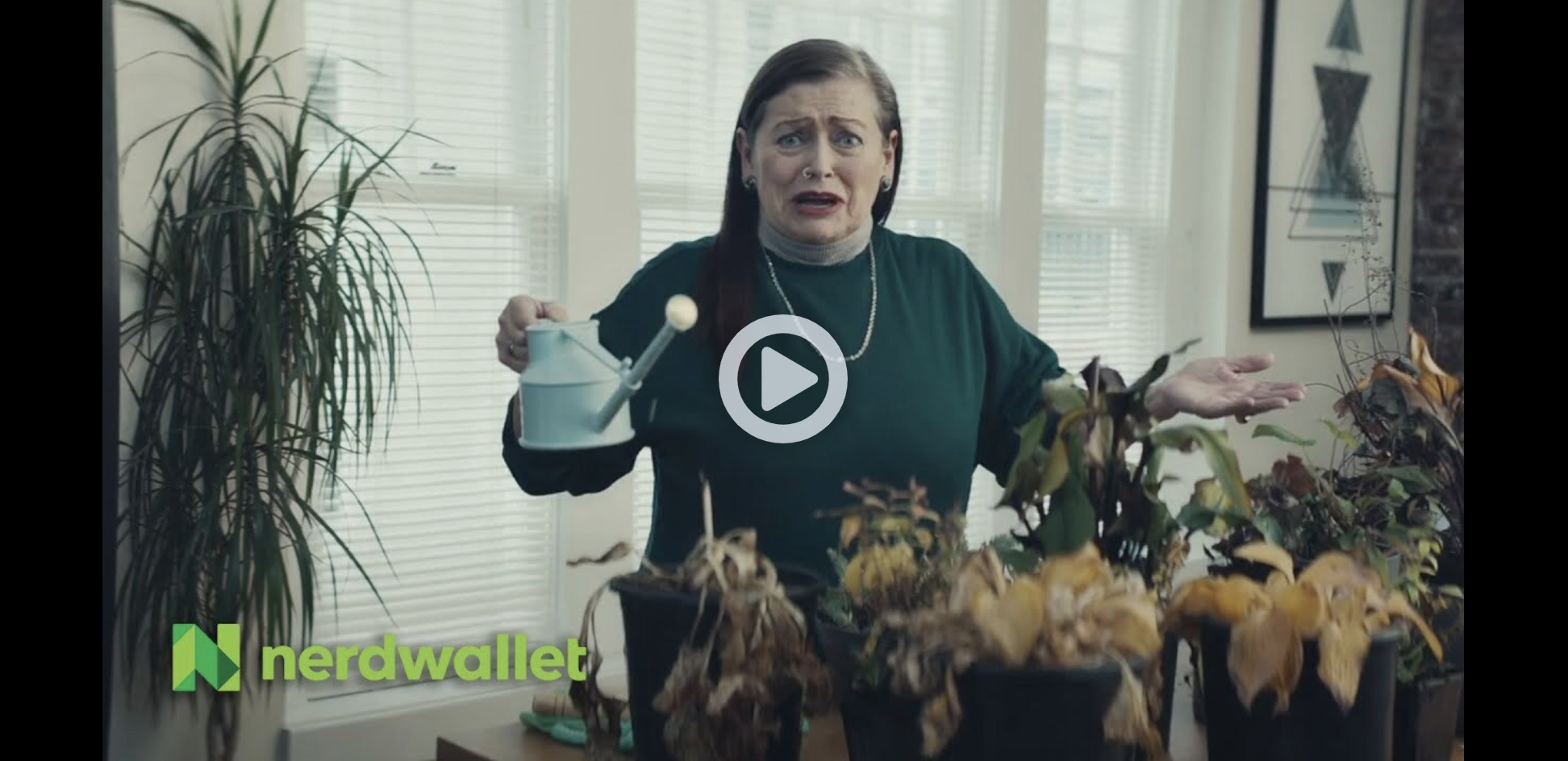 A woman with brown hair, wearing a green top and a pearl necklace, appears distressed while holding a small watering can and standing behind a table with withered potted plants. Behind her are large windows with blinds, a tall plant, and framed geometric art on the wall.