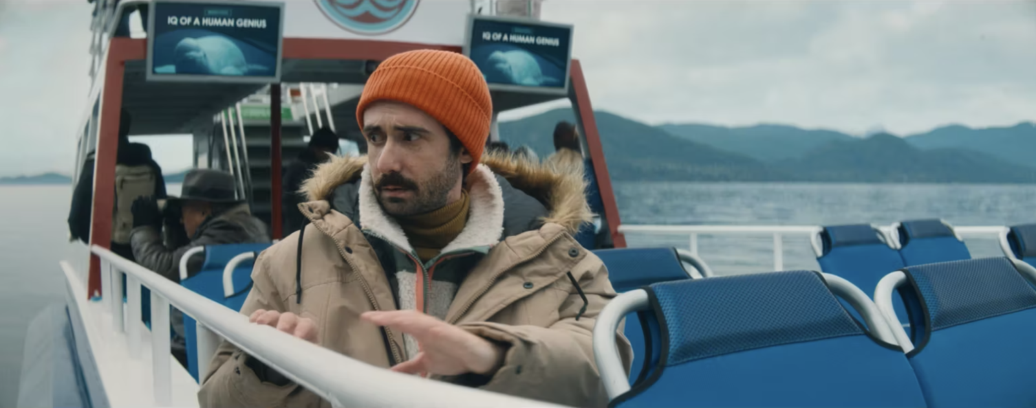 A man with a beard wearing an orange beanie and a tan jacket with a fur-lined hood sits on a boat near a body of water, with a mountainous landscape in the background. He appears to be looking to his left with a concerned expression.