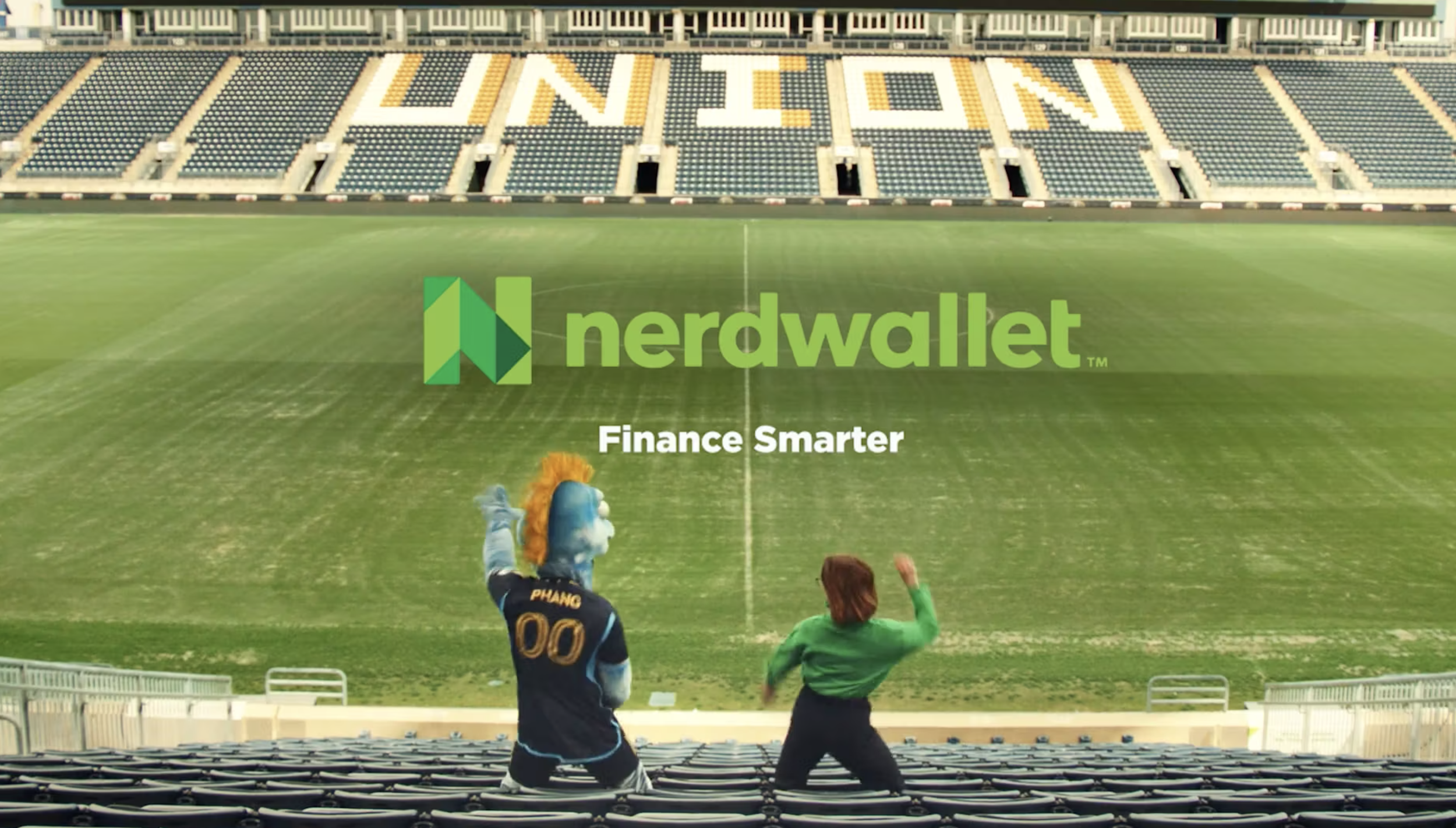 A football stadium with the word 'UNION' displayed on the seats in the background. Two mascots, one with an orange mane and blue face marked 'Phang 00' and the other in green, are in the foreground, seated on the seats and facing the field. The field has grass markings.