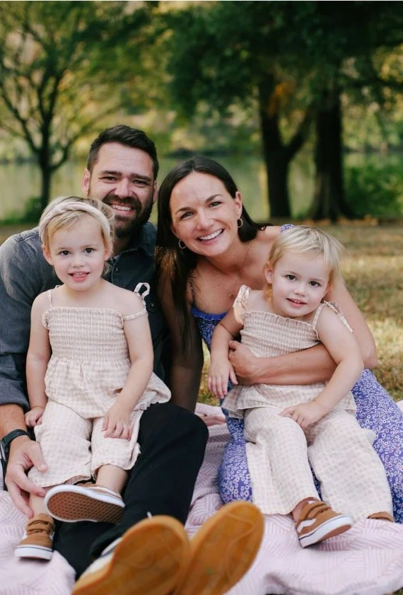Family of four sitting outdoors on a blanket in a park, smiling at the camera. The group includes two adults and two young girls, all dressed in casual summer clothing, with trees in the background.