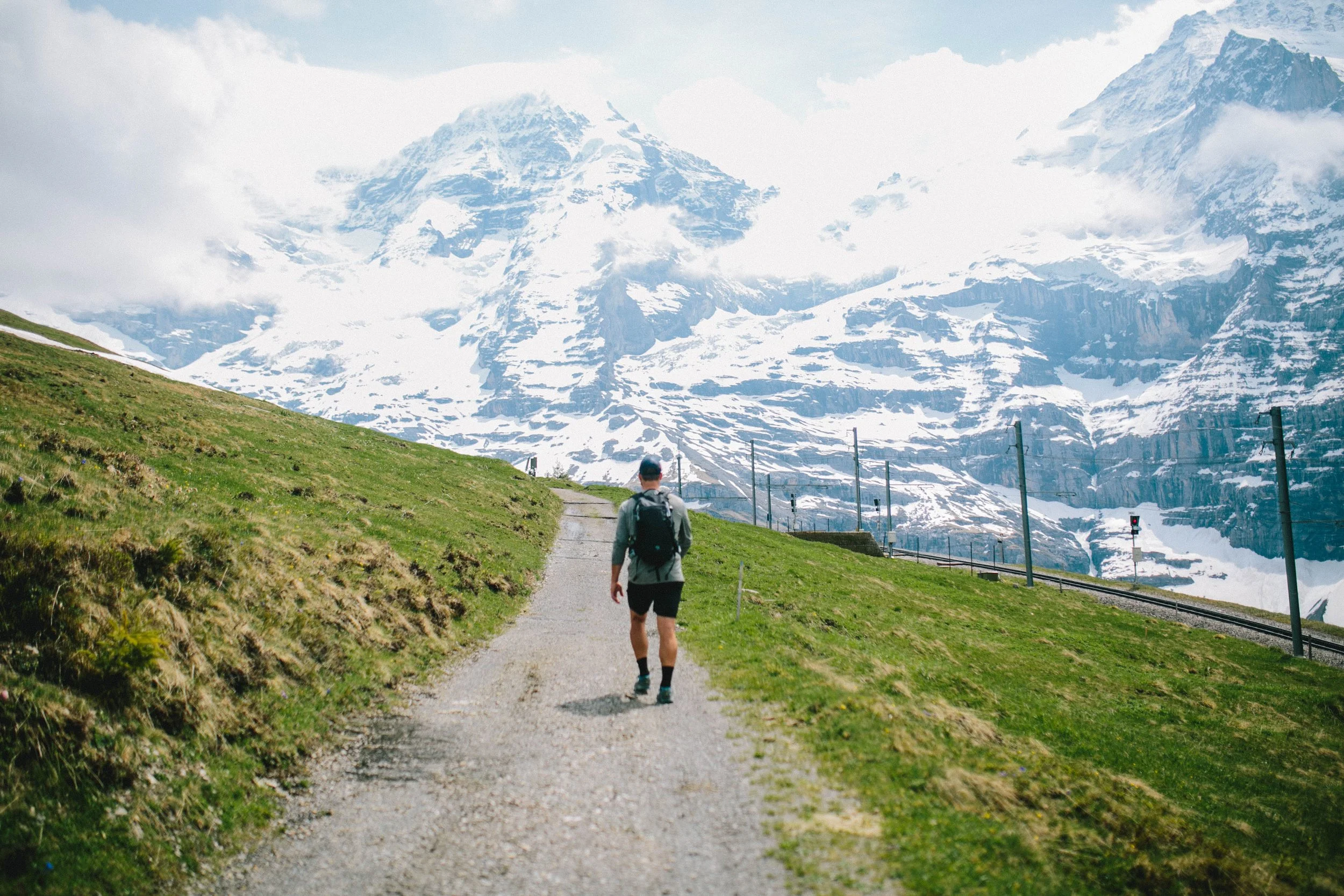 A hiker with a backpack walking on a dirt trail through green grass, with snow-capped mountains in the background.