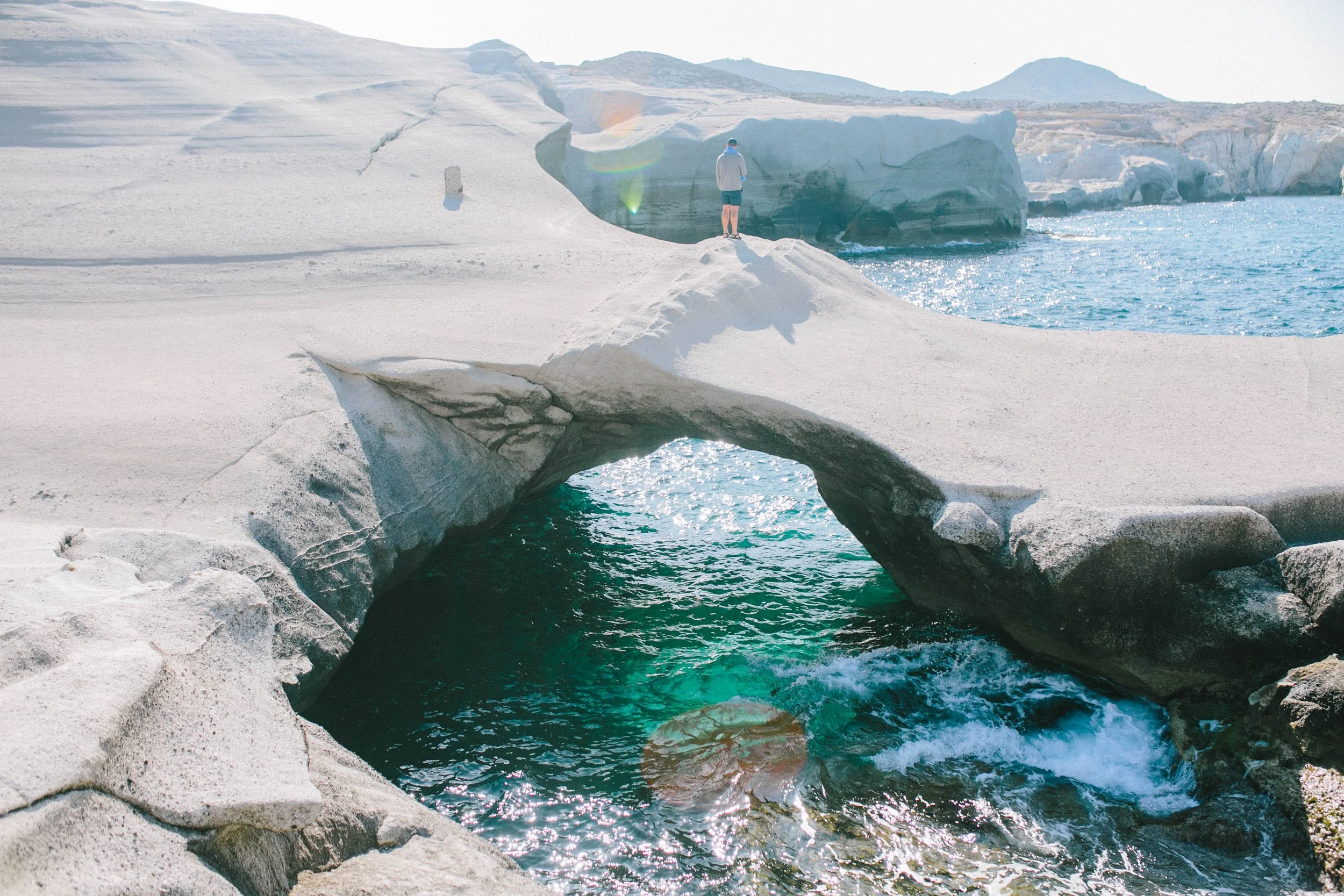 A person standing on a natural rock arch over a turquoise sea, with rocky white landscape and icebergs in the background on a bright sunny day.