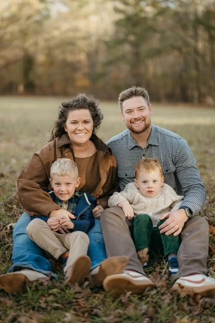 A family of four sitting on the grass outdoors during fall, with trees in the background. The woman and man are smiling, and the two young boys sit on their laps, also smiling.