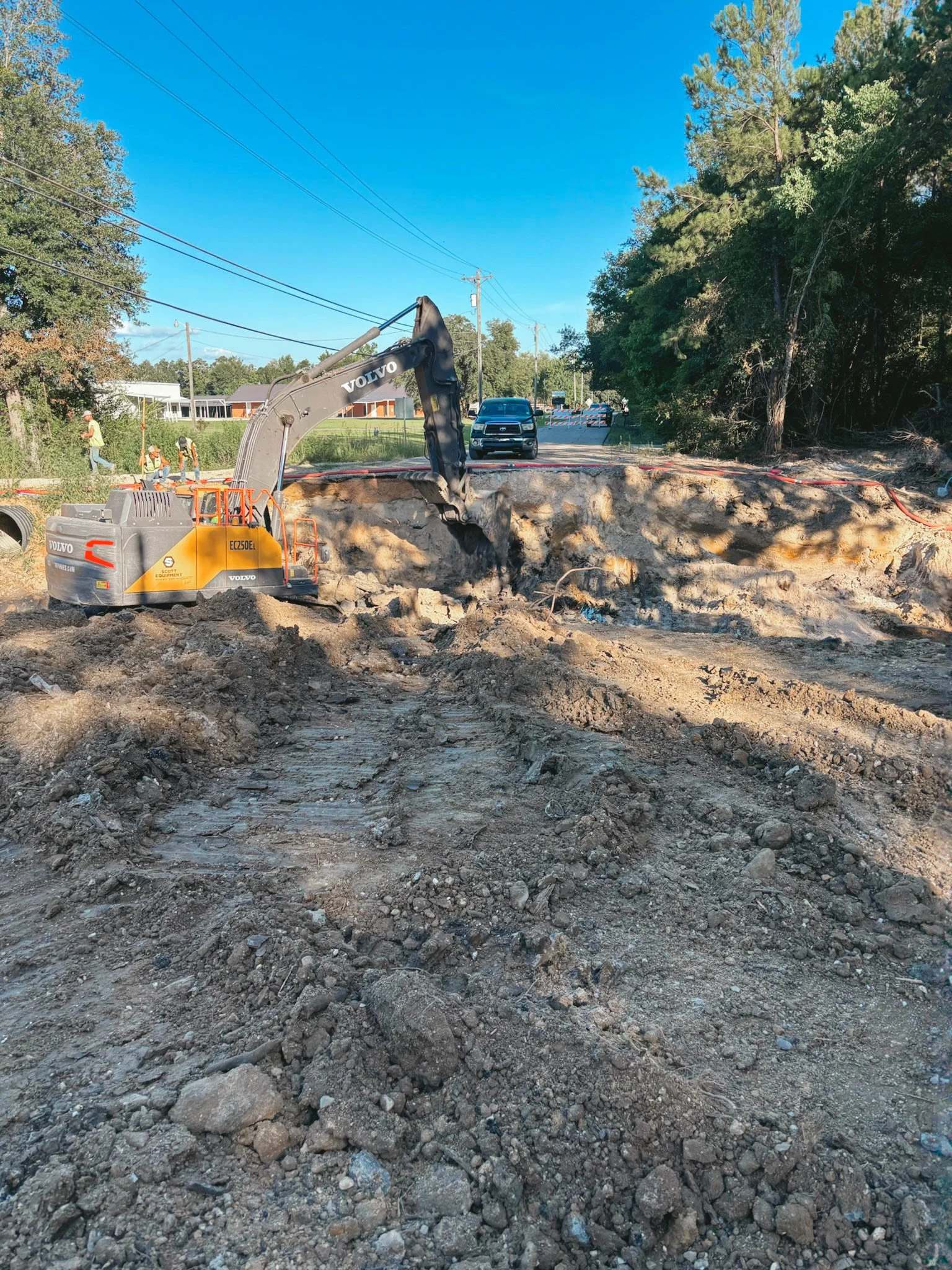 Construction site with an excavator scoop in dirt and a car in the background, with trees and power lines.