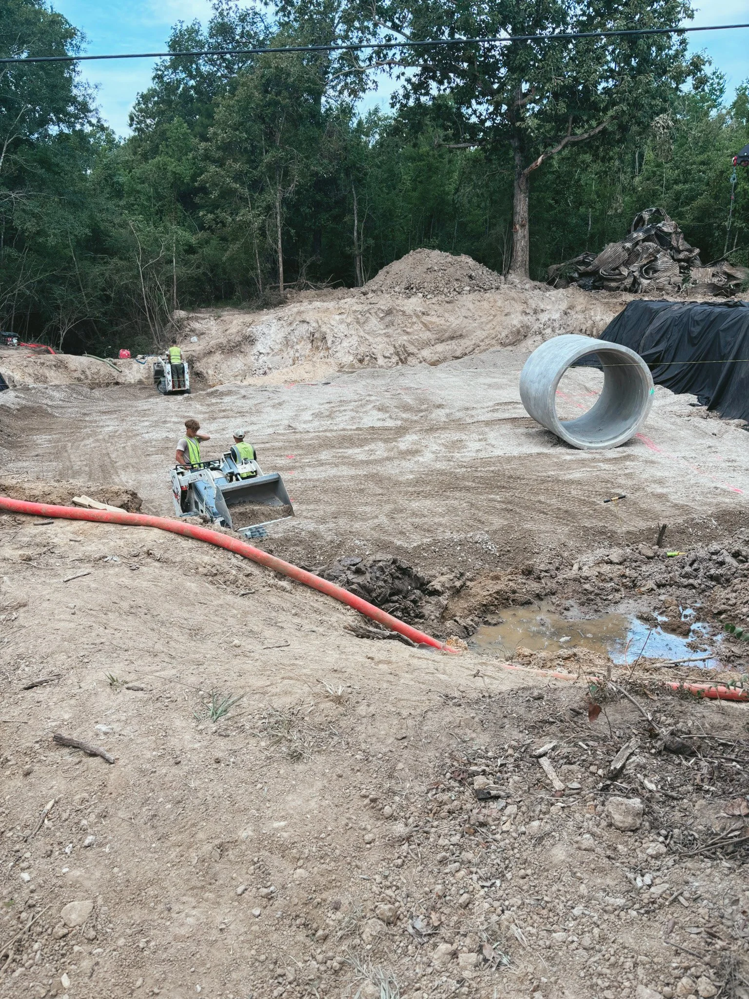 Construction site with workers operating machinery, large concrete pipe, and gravel pile in a wooded area.