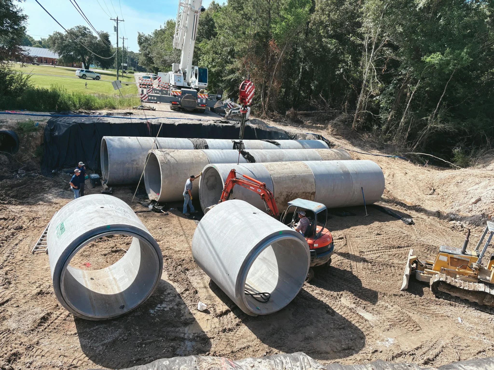 Construction workers installing large concrete pipes in a trench for infrastructure work, with a crane and excavator present, and trees in the background.