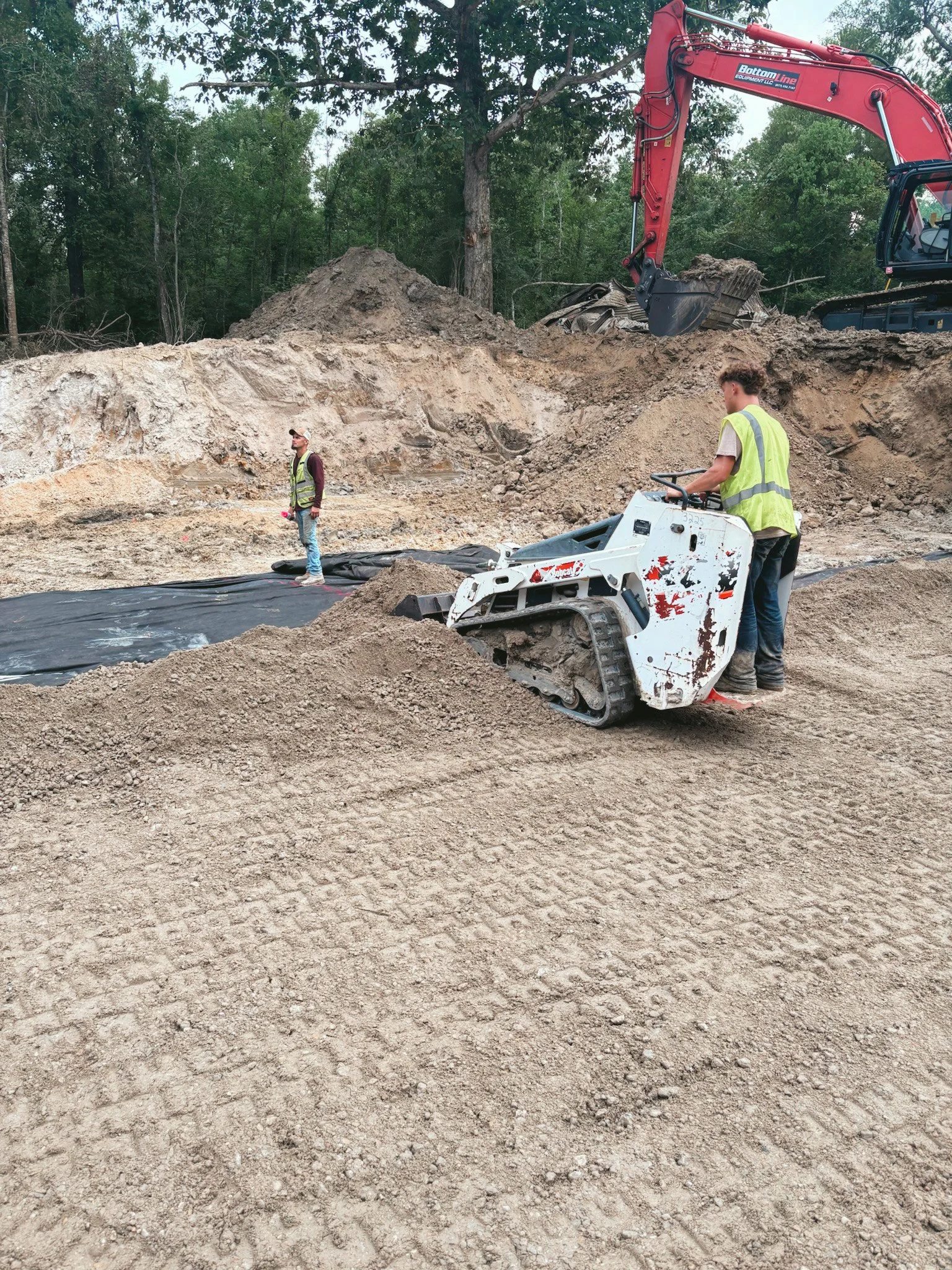 Construction site with two workers, one operating a compact skid steer loader and the other standing nearby, with a large excavator in the background, surrounded by dirt and trees.
