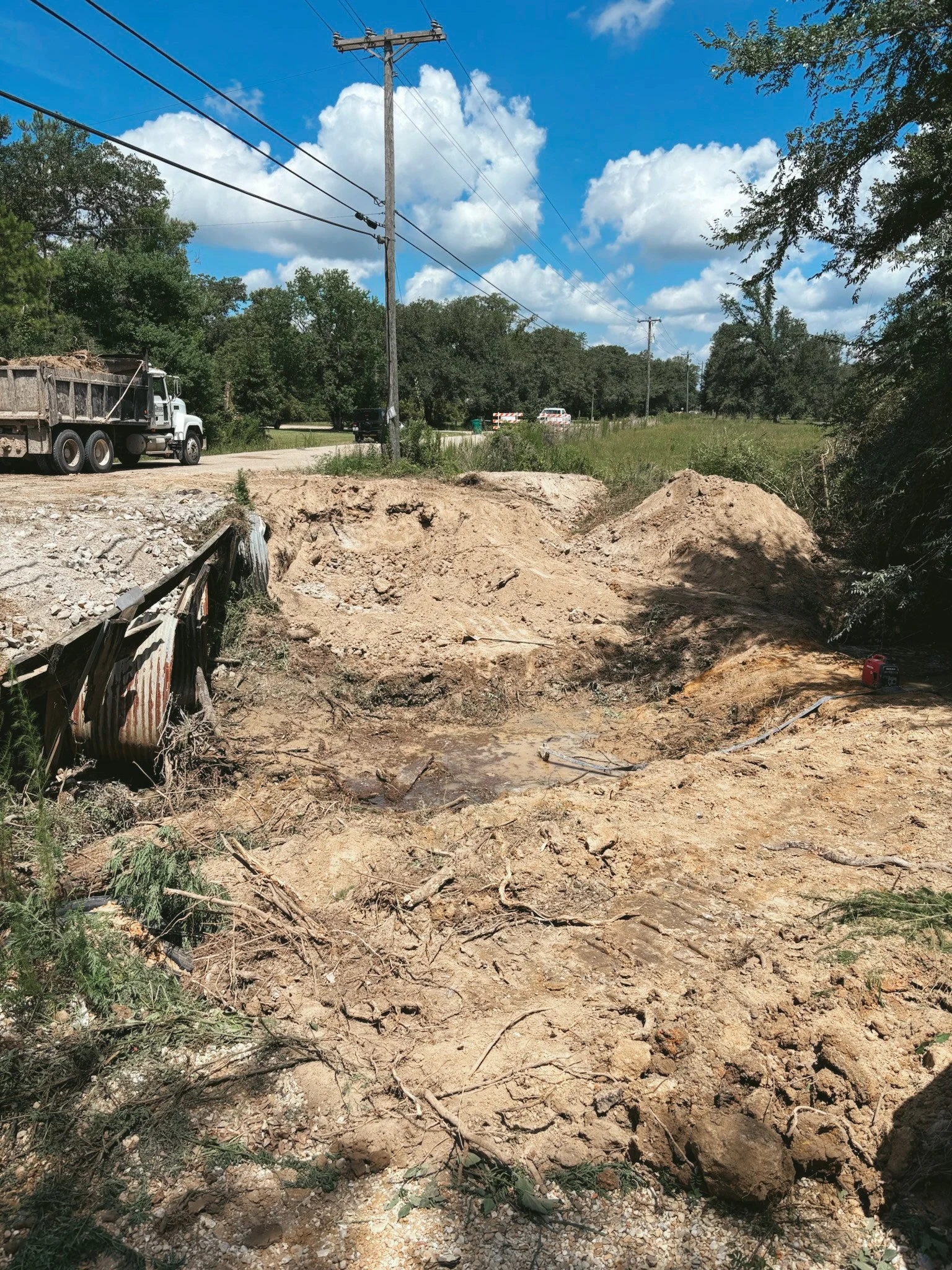 A construction site on a roadside with disturbed earth, dirt mounds, and exposed pipe, with utility poles and trees in the background under a bright blue sky with clouds.