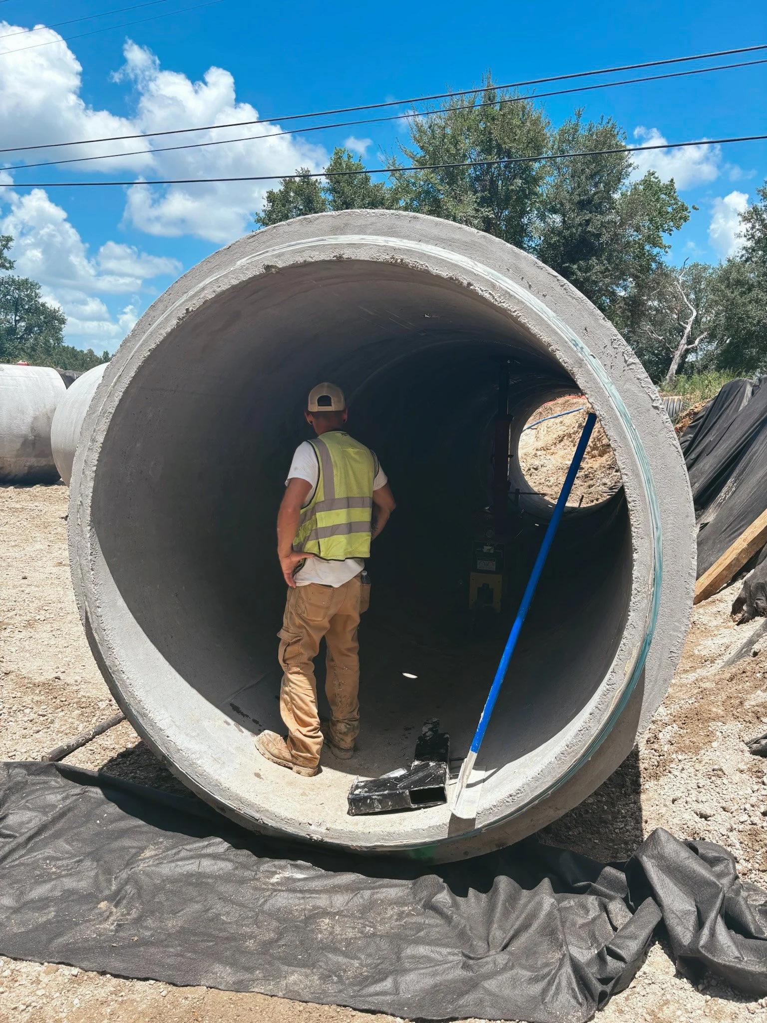 A construction worker in a safety vest and cap standing inside a large concrete pipe at a construction site, with a blue sky and some trees in the background.