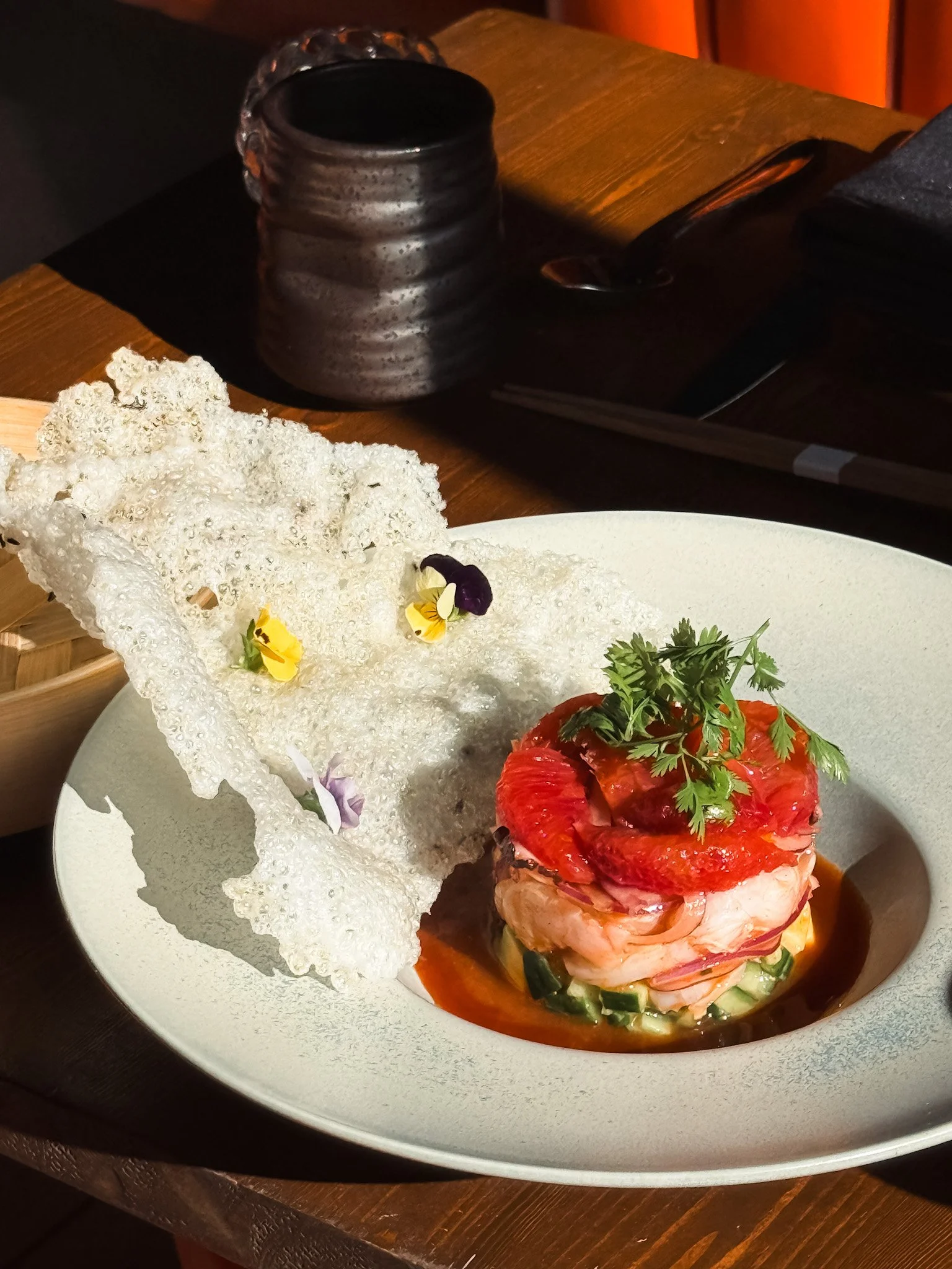 A gourmet dish featuring a stack of sliced tomatoes and seafood, garnished with cilantro, alongside a decorative rice cracker topped with edible flowers, all served on a white plate.