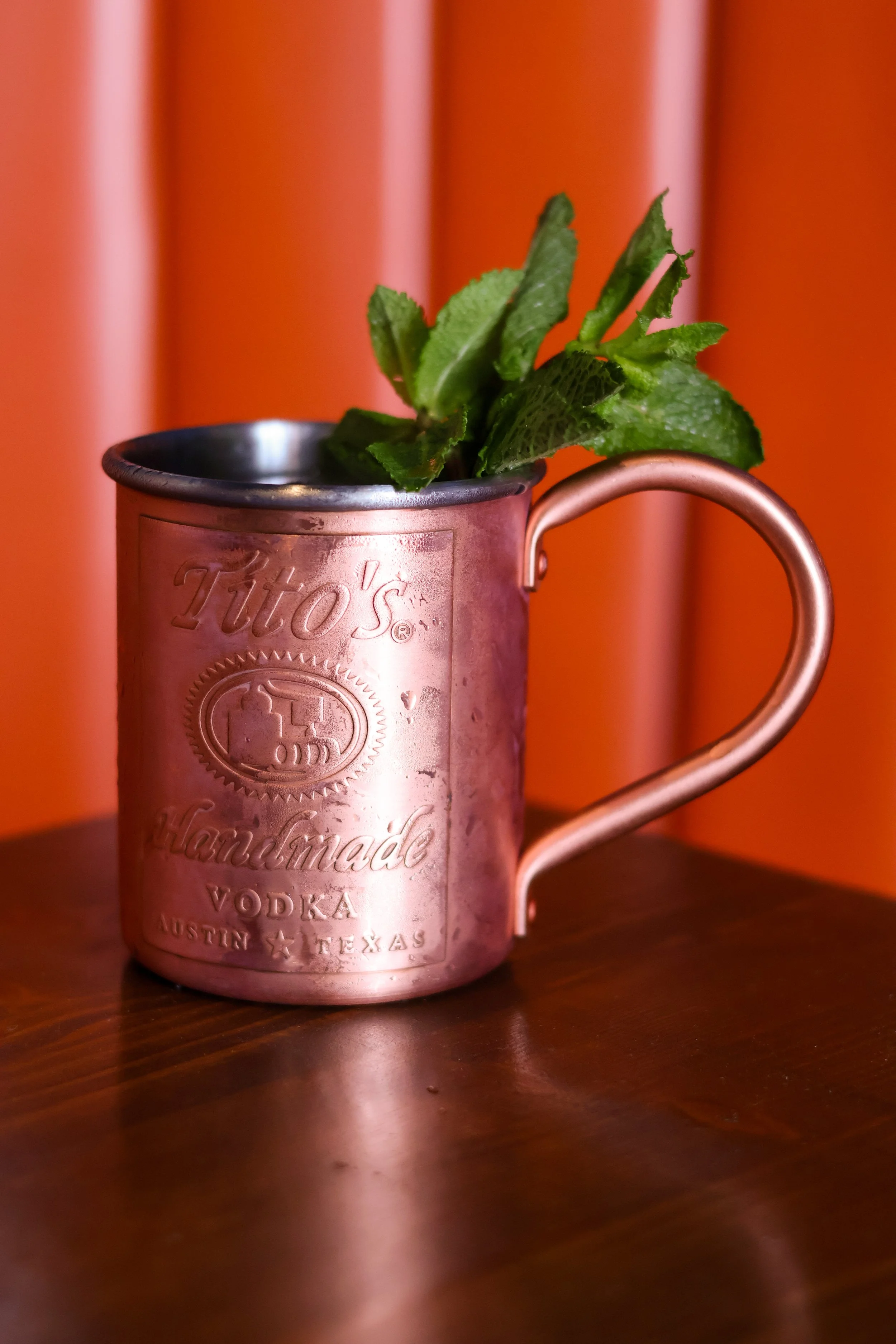 A copper mug with an embossed logo, containing fresh mint leaves, placed on a wooden surface with a warm-colored background.
