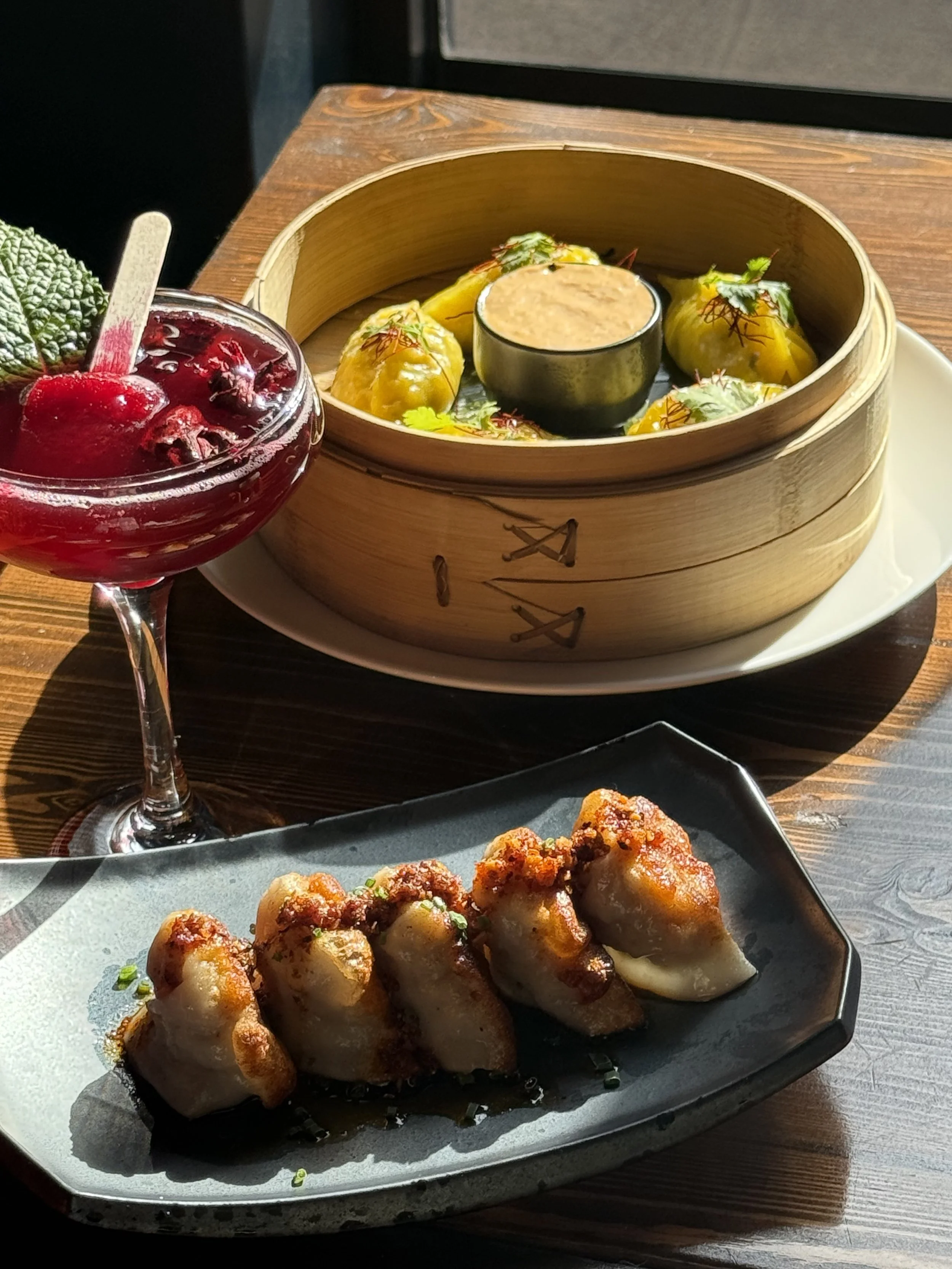 A plate of five dumplings with sauce on a black rectangular dish, a bamboo steamer containing more dumplings and dipping sauce, and a cocktail garnished with mint, cherry, and berry atop a wooden table.