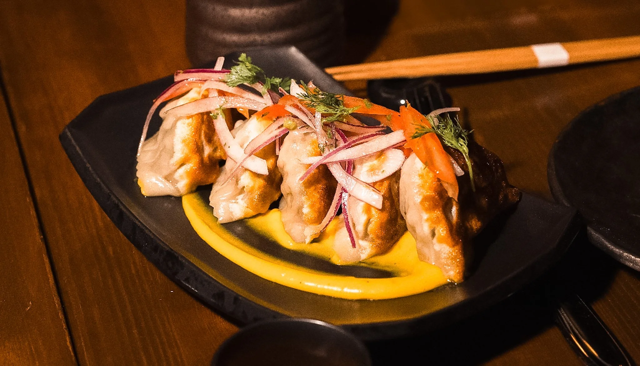 Plate of four Asian dumplings topped with shredded onions, carrots, and herbs, served on a black plate with a yellow sauce underneath.