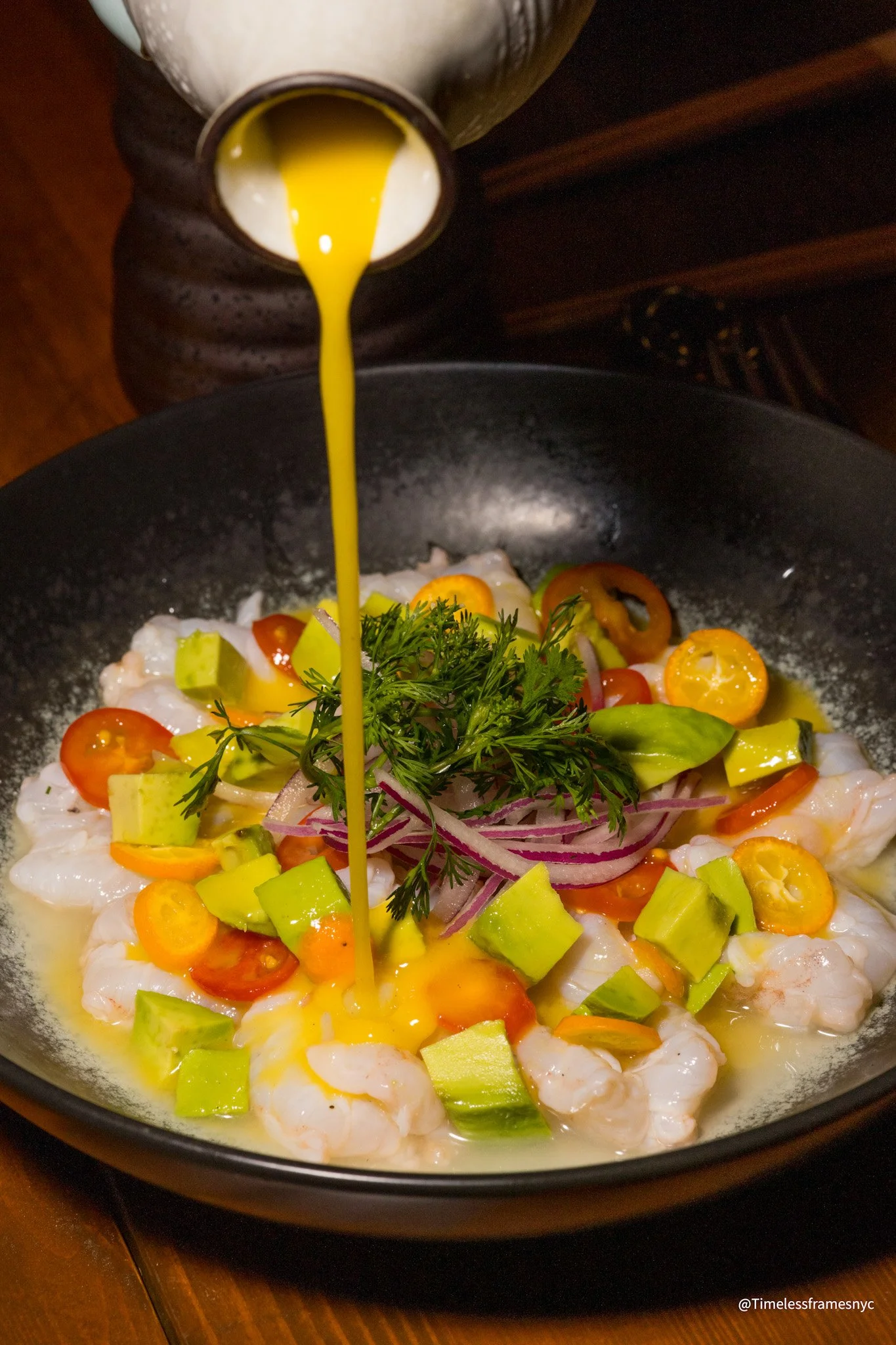 Pouring yellow sauce over a bowl of shrimp, cherry tomatoes, avocado, and herbs