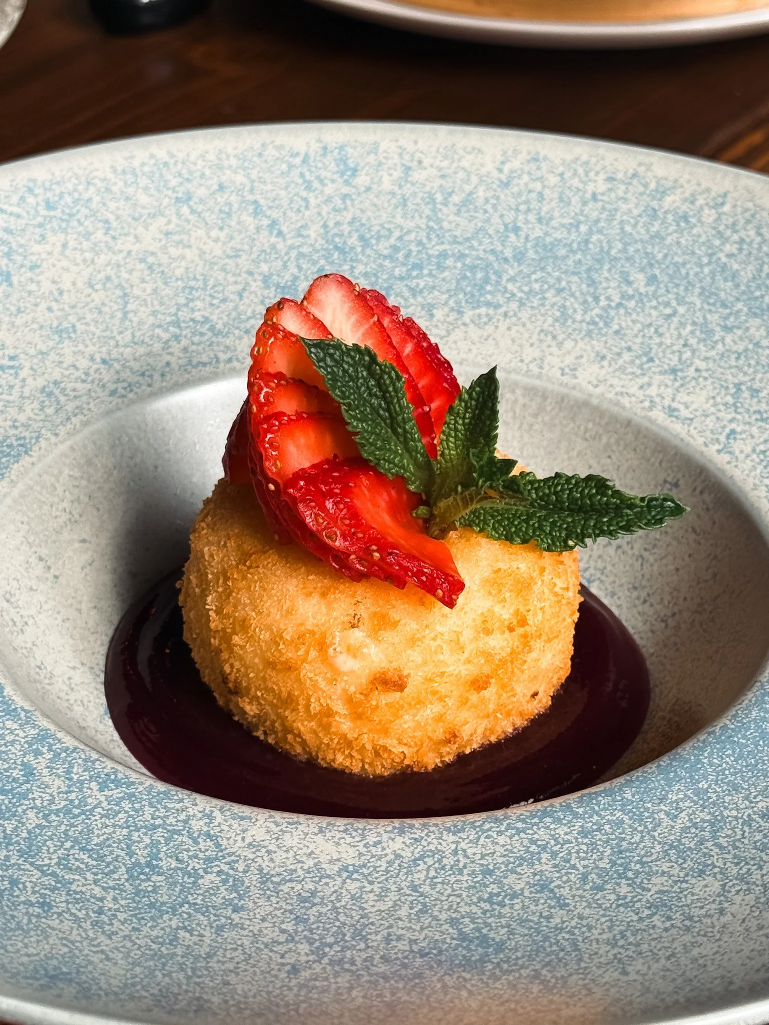 A breaded and fried ball dessert topped with sliced strawberries and a sprig of mint, served on a dark sauce in a white bowl with blue speckles.