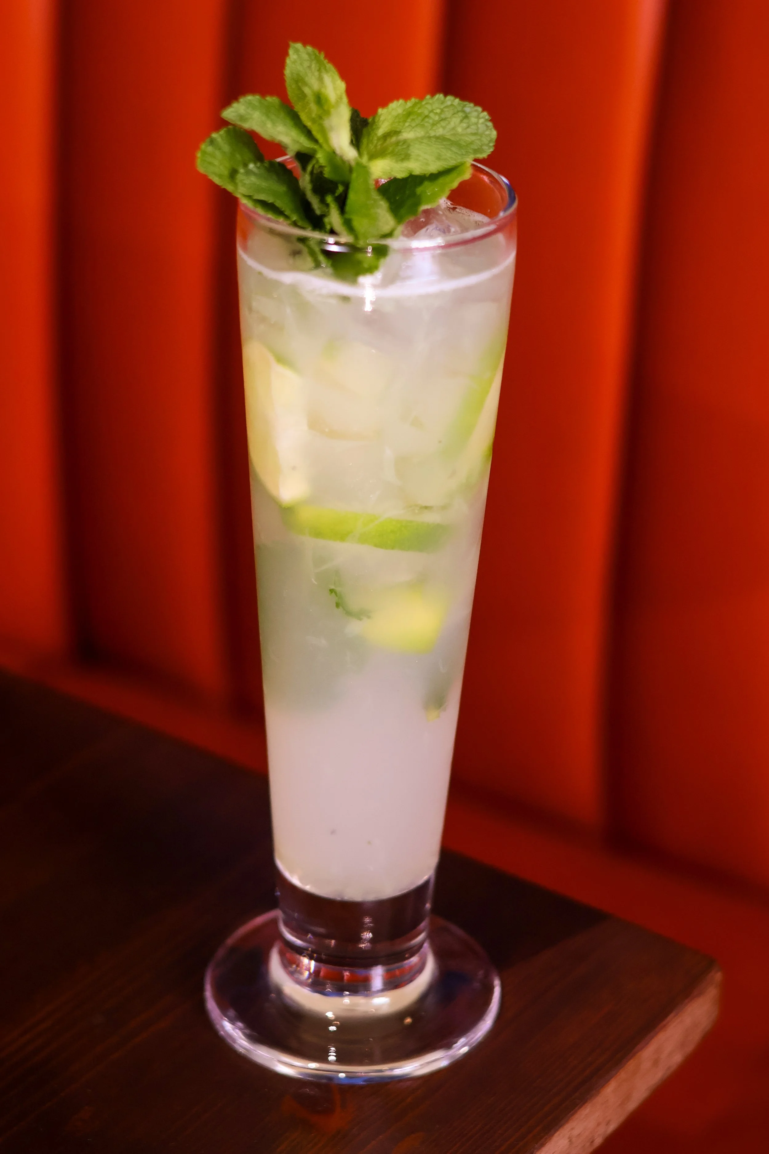 A tall glass of mojito cocktail with lime slices and fresh mint leaves garnish, placed on a wooden surface against a red background.
