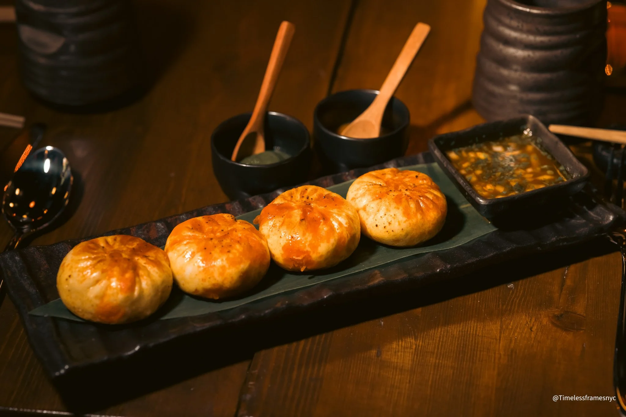 Four cooked Asian-style dumplings on a rectangular black plate with dipping sauces and condiments in black bowls in the background.