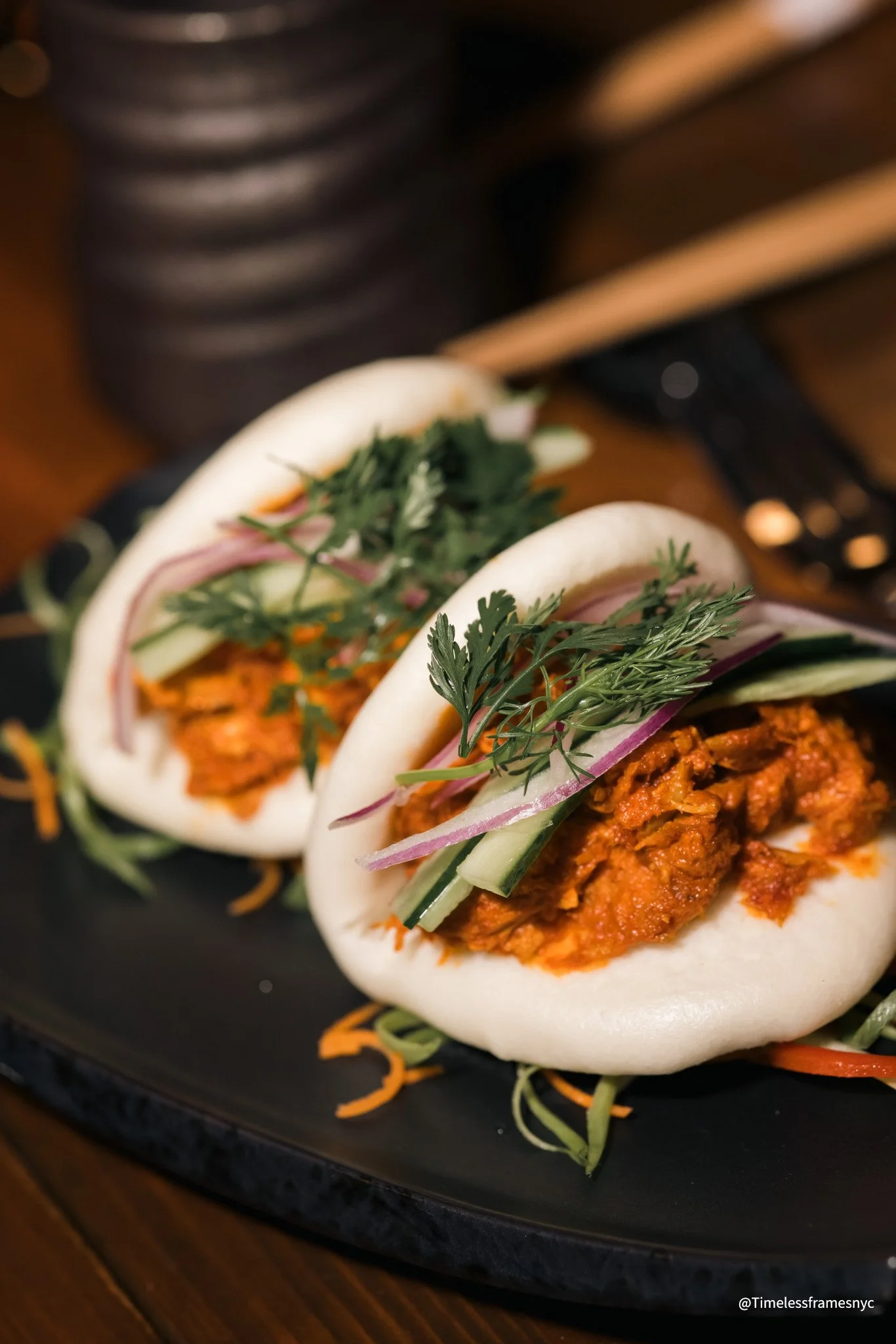 Two steamed bao buns filled with spiced meat and topped with cilantro, cucumber, red onion, and shredded carrots, served on a black plate.