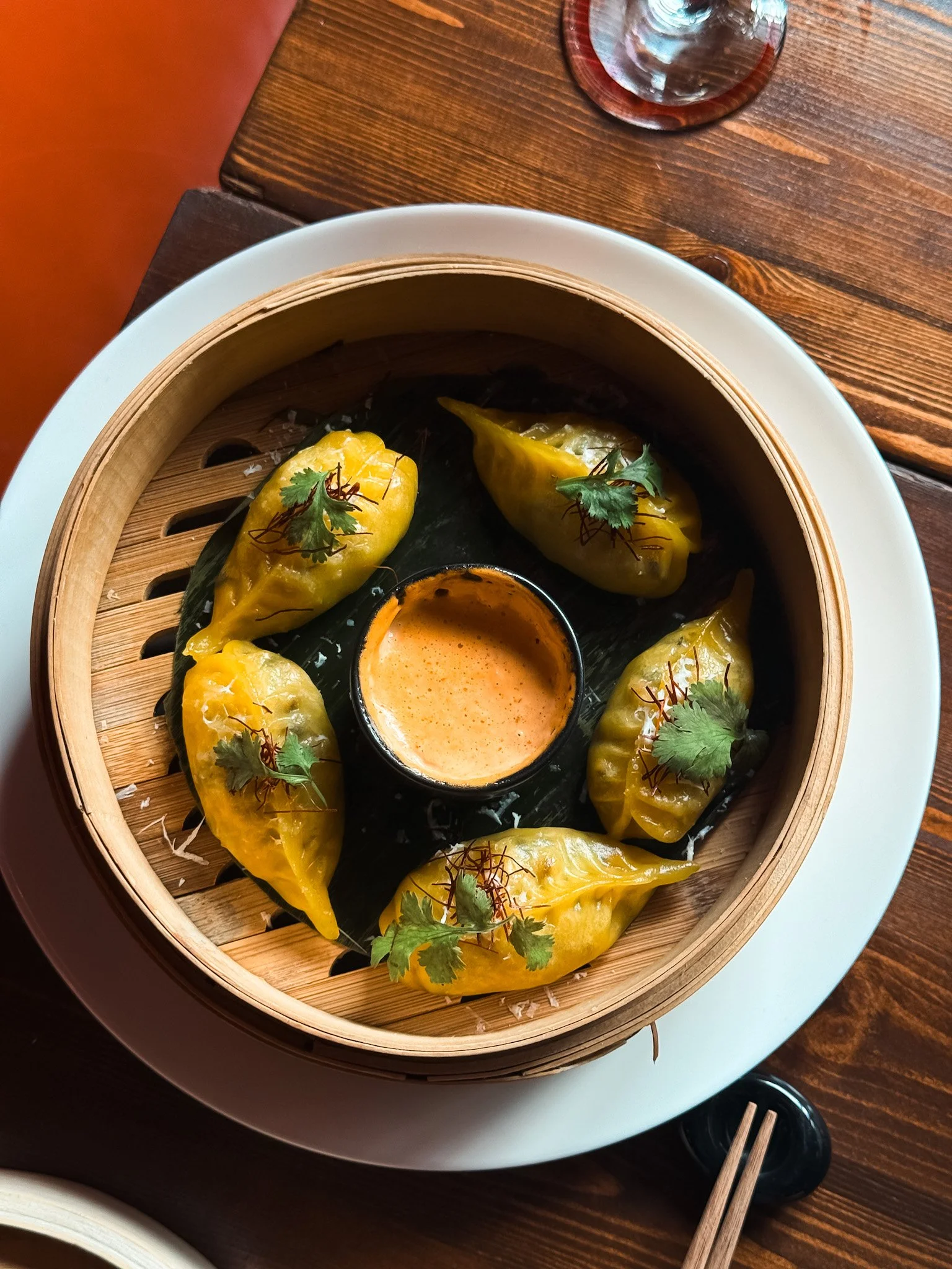 Six yellow dumplings garnished with cilantro leaves arranged in a circle inside a bamboo steamer on a white plate, with a small cup of orange dipping sauce in the center.