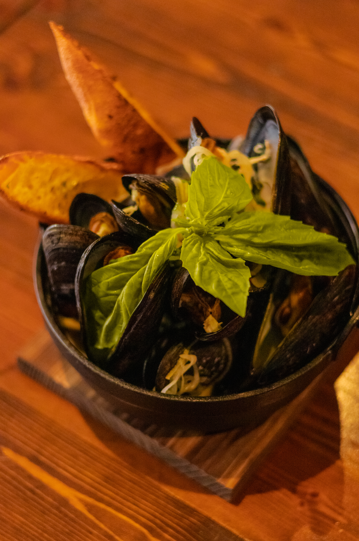 A bowl of cooked mussels garnished with fresh basil leaves and served with toasted bread on the side.