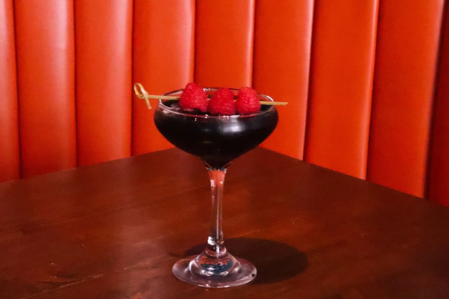 Dark berry cocktail garnished with raspberries in front of an orange leather banquette