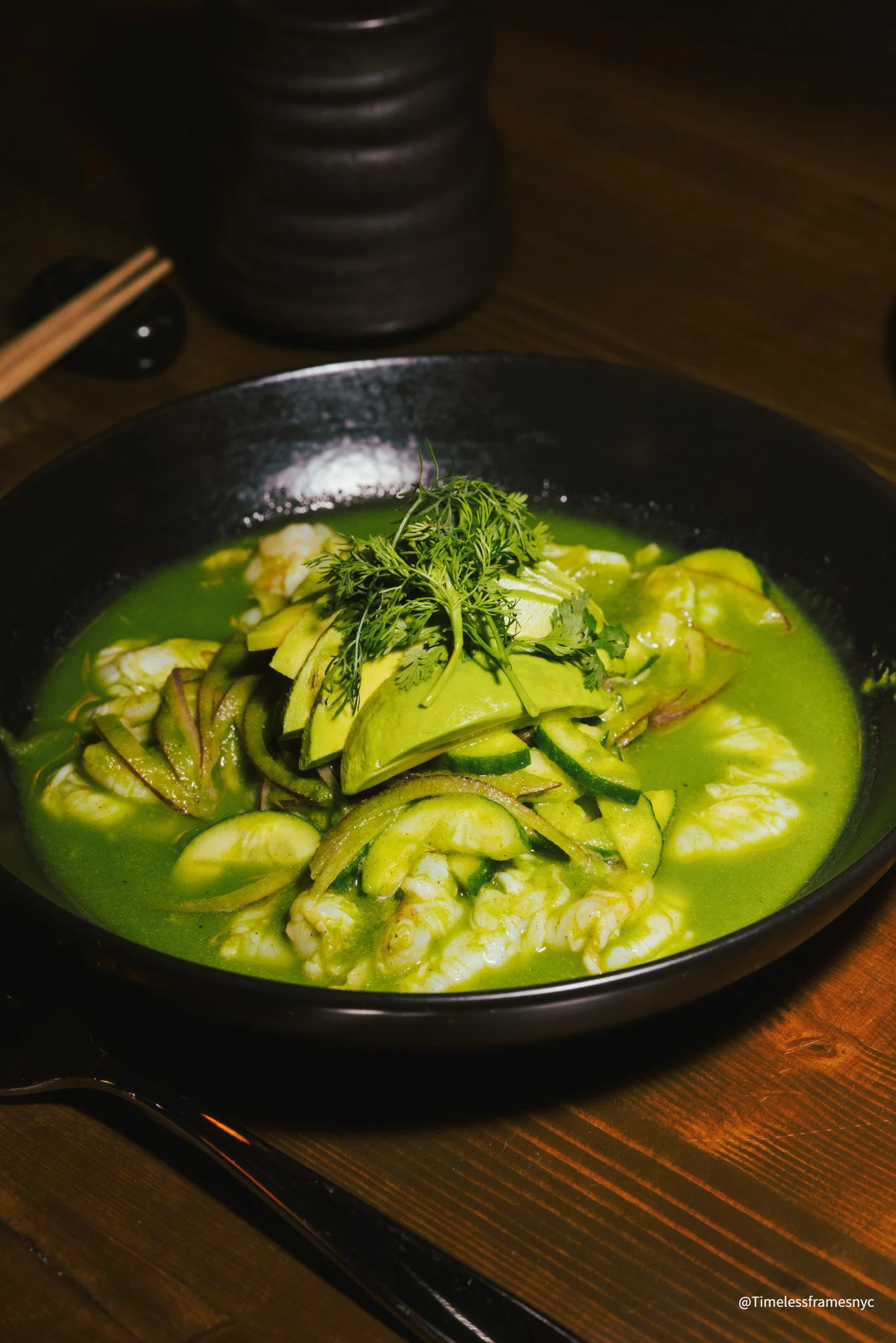 A bowl of green-colored soup garnished with sliced avocado, fresh herbs, and possibly zucchini, served on a dark wooden table.