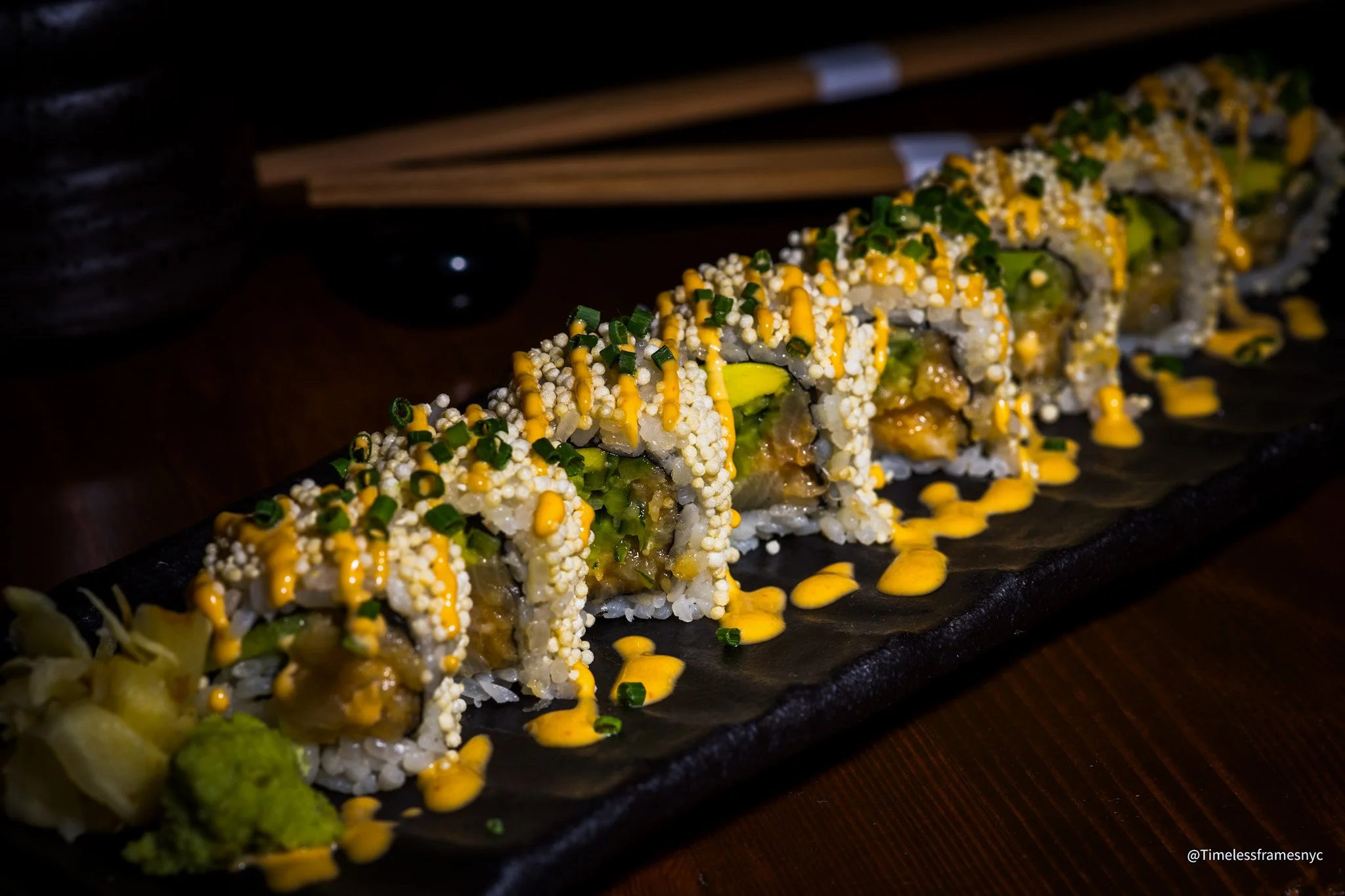 Sushi roll with rice, seaweed, avocado, cooked fish, topped with chopped chives and orange sauce, served with pickled ginger and wasabi on the side.